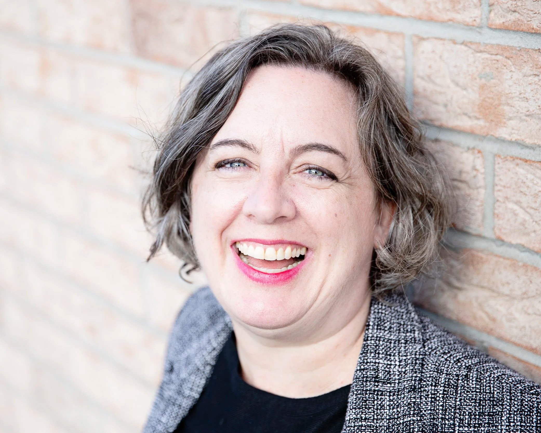 Close-up of a woman with gray hair smiling and winking, standing against a brick wall.