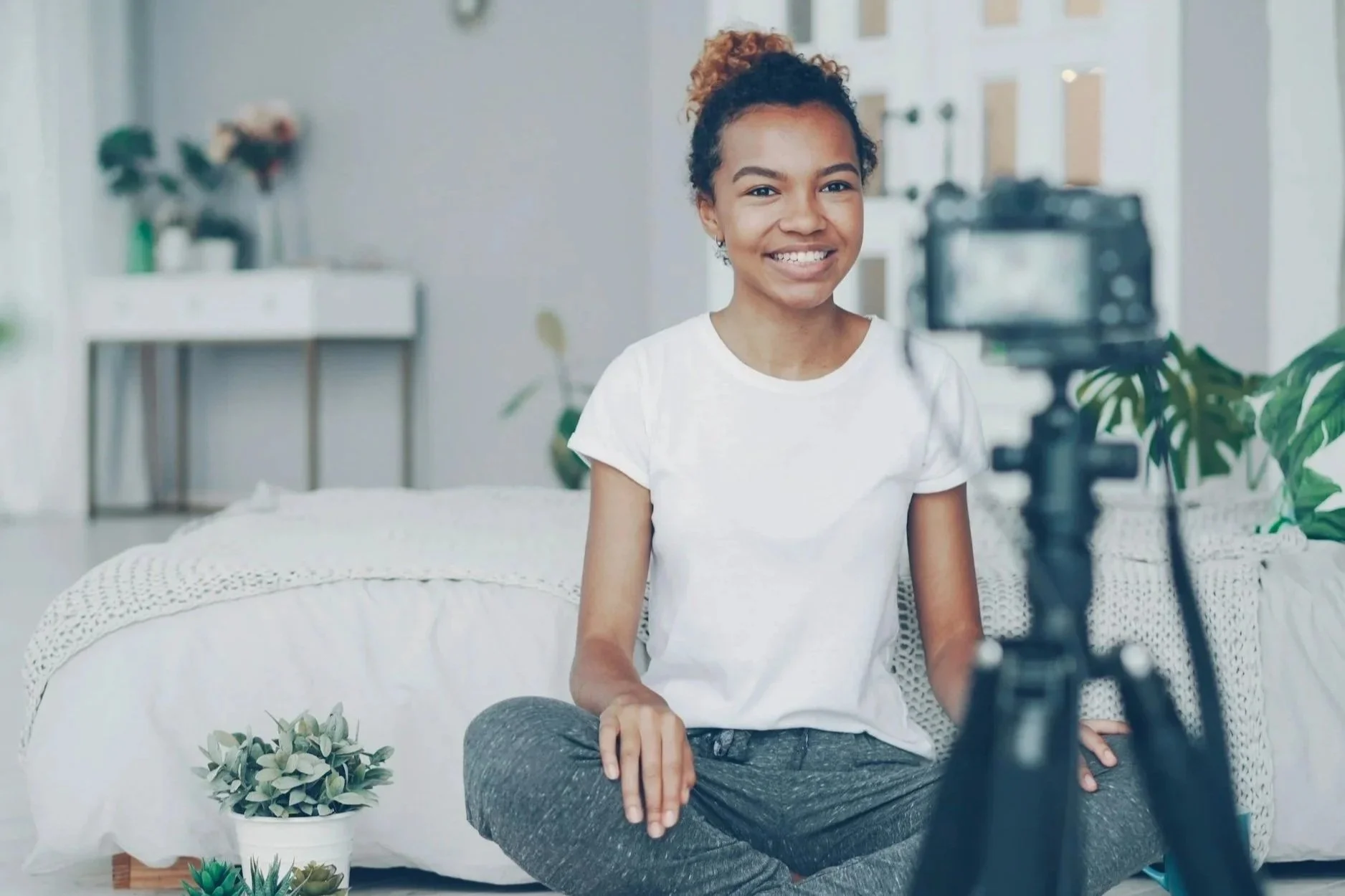 Young woman with curly hair in a bun, sitting cross-legged on a bed, smiling at a camera on a tripod, in a well-lit bedroom with plants and furnishings.