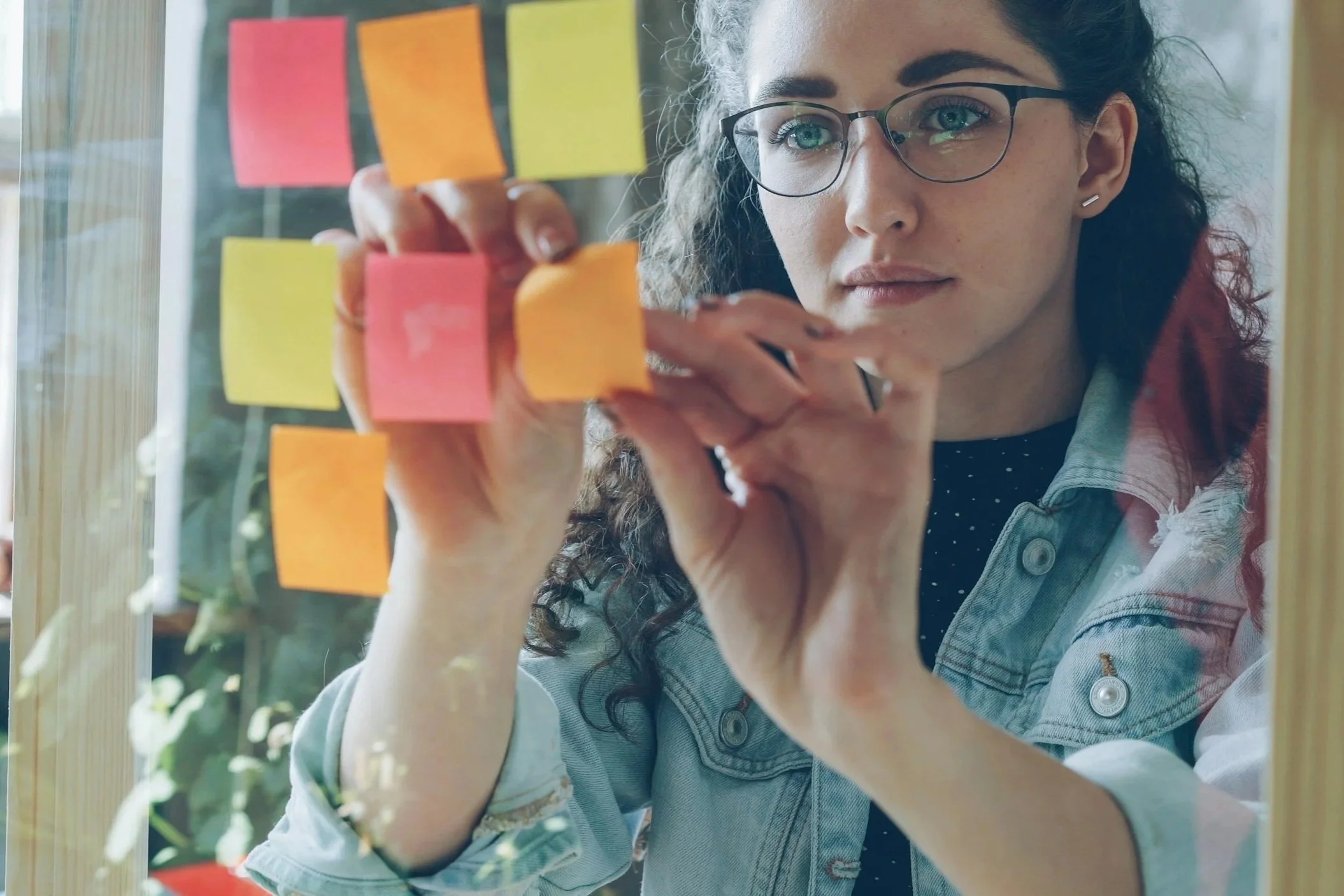 A woman with curly hair, glasses, and earrings, drawing pink, yellow, and orange sticky notes on a glass wall or window.