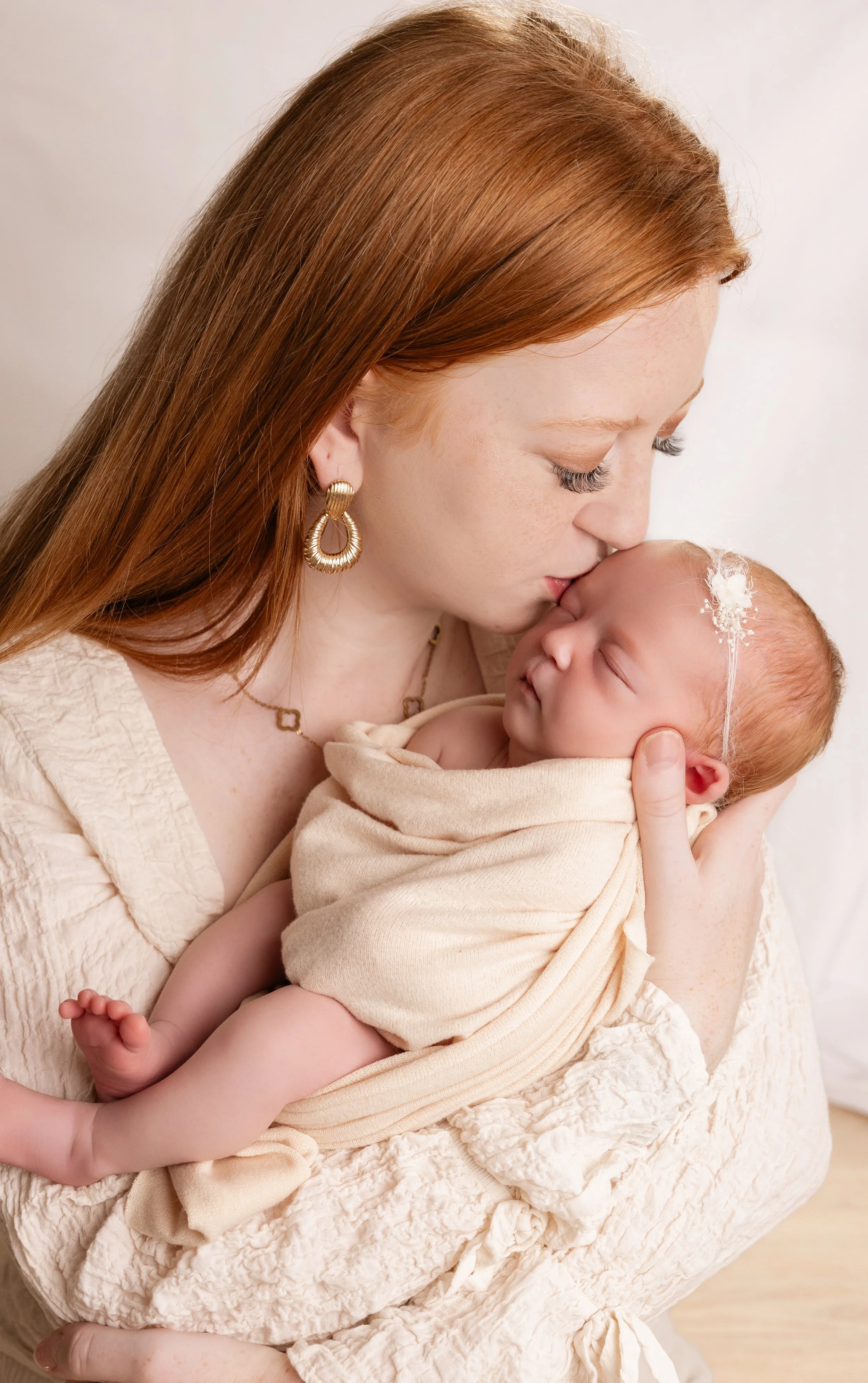 Une femme aux cheveux roux tient un bébé blotti contre elle, lui donne un baiser sur la tête dans un cadre intérieur avec lumière douce.