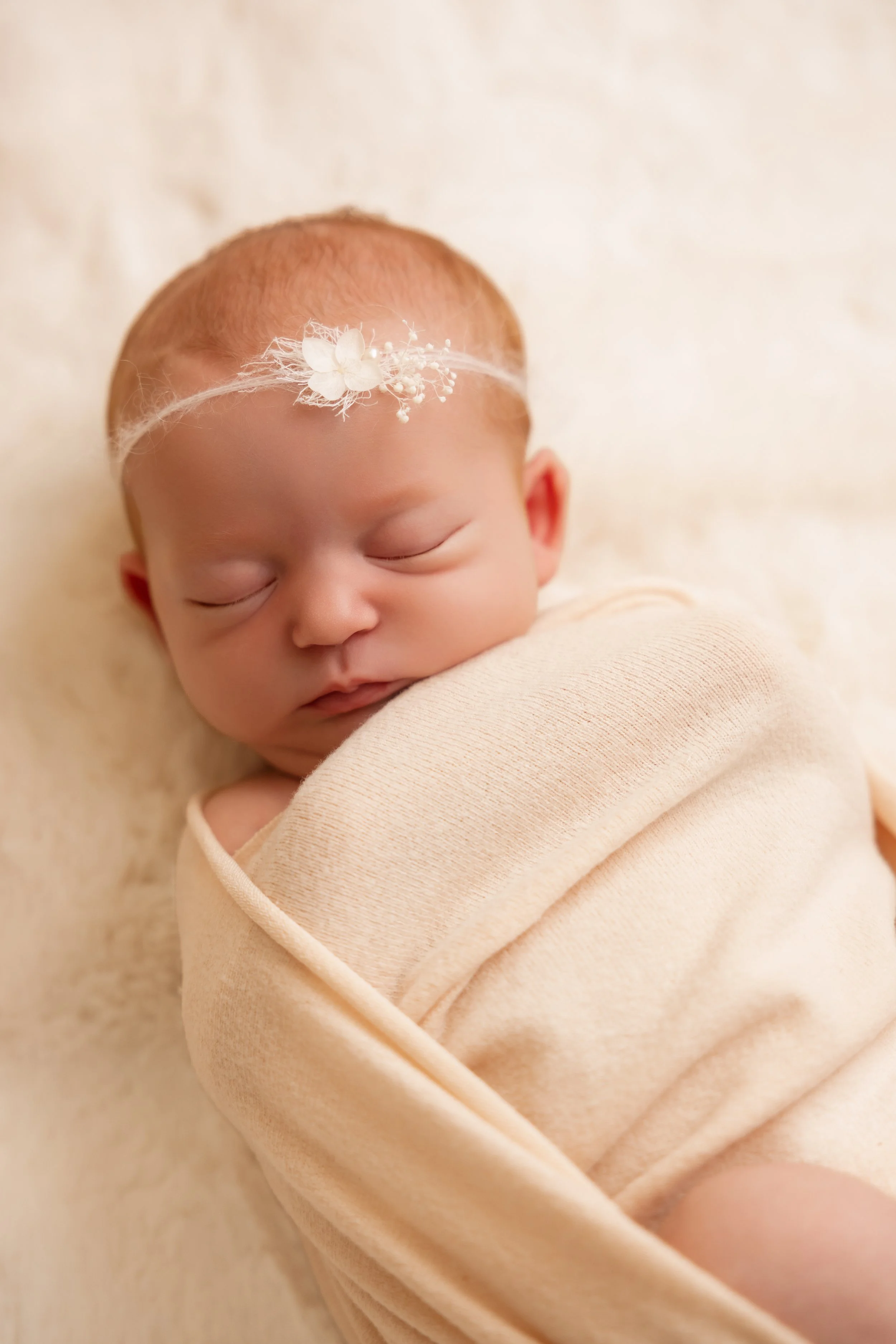 Un bébé nouveau-né endormi, enveloppé dans une couverture beige claire, portant une bandeau blanc avec une petite décoration florale.