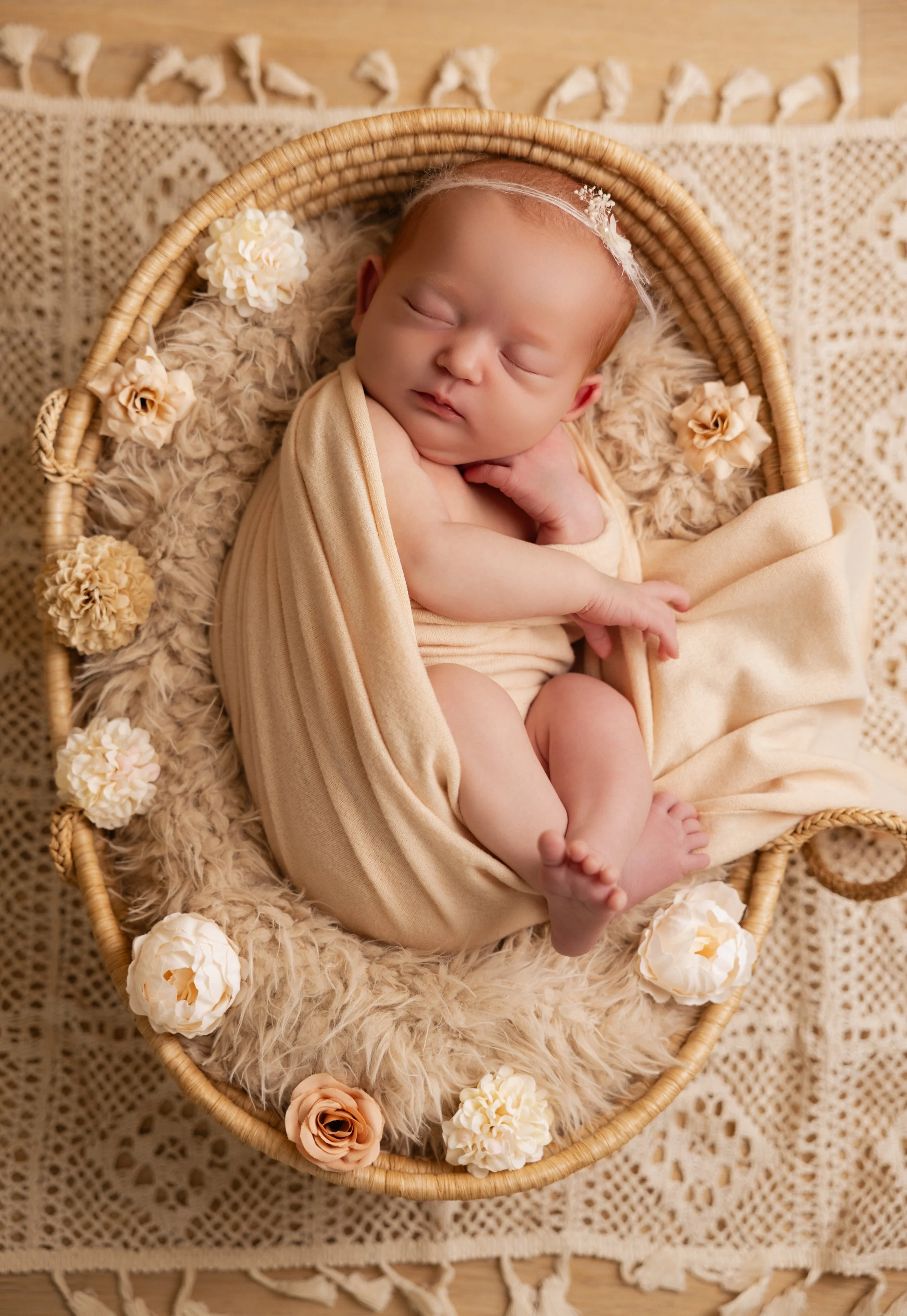 Un bébé endormi dans un panier en rotin beige, entouré de fleurs en tissu blanches, ivoire et pêche, sur un matelas en fausse fourrure beige, posé sur une nappe crochet beige.