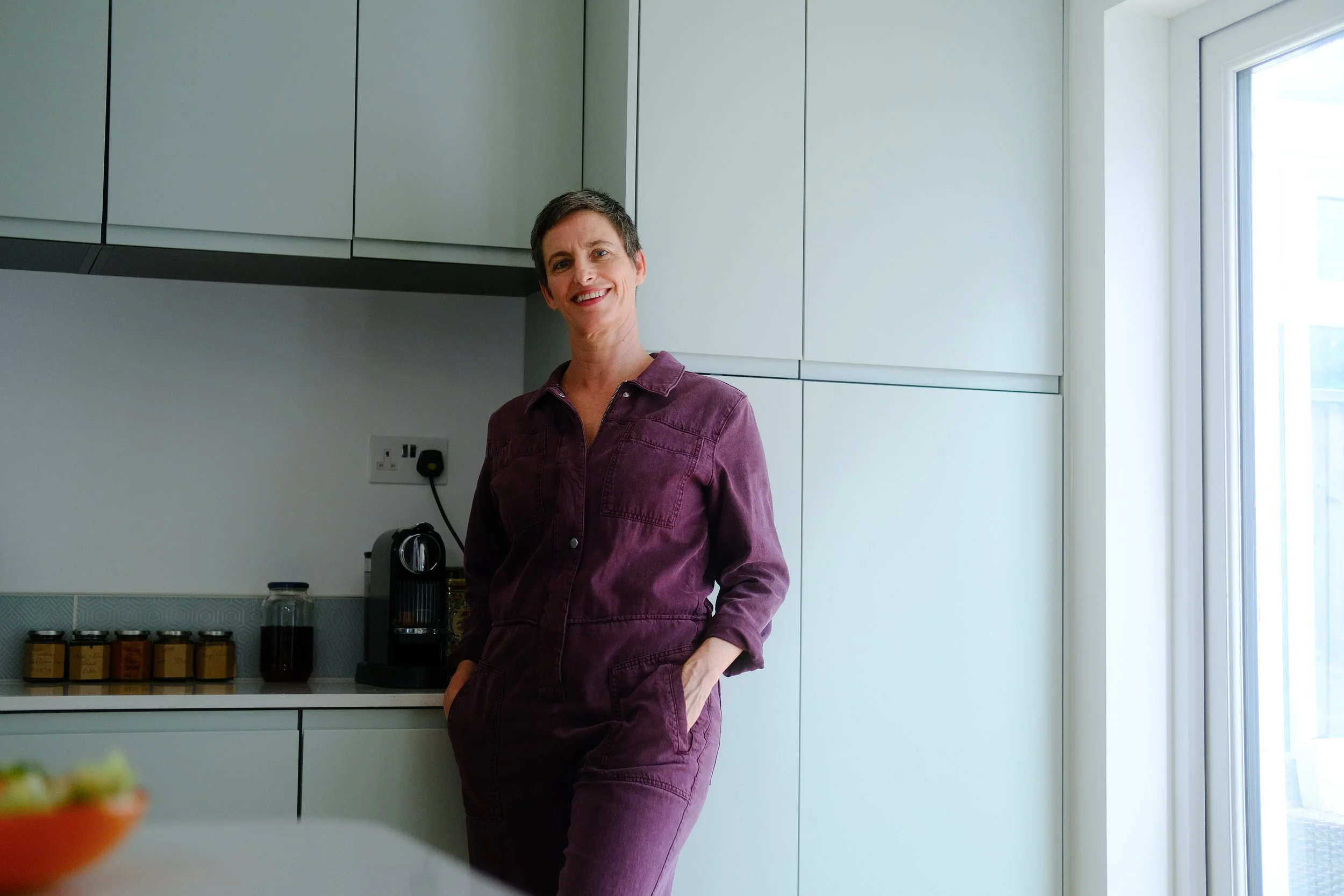 A woman with short dark hair smiling in a modern kitchen, wearing a maroon jumpsuit, standing near a window, with kitchen cabinets and appliances visible in the background.