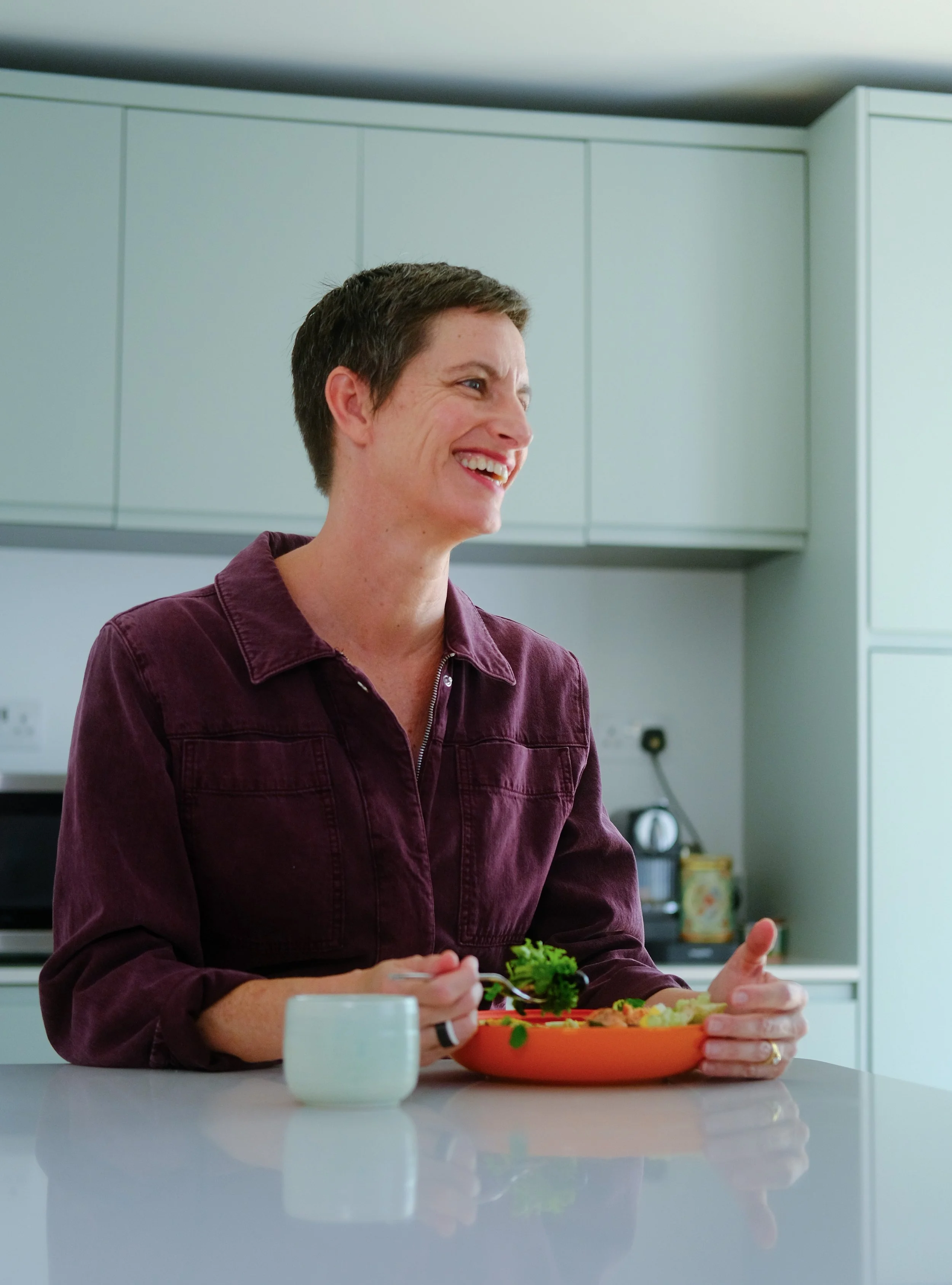 A woman with short dark hair and a maroon jumpsuit smiling while holding a fork and a small bowl with green leafy vegetables in a modern kitchen.