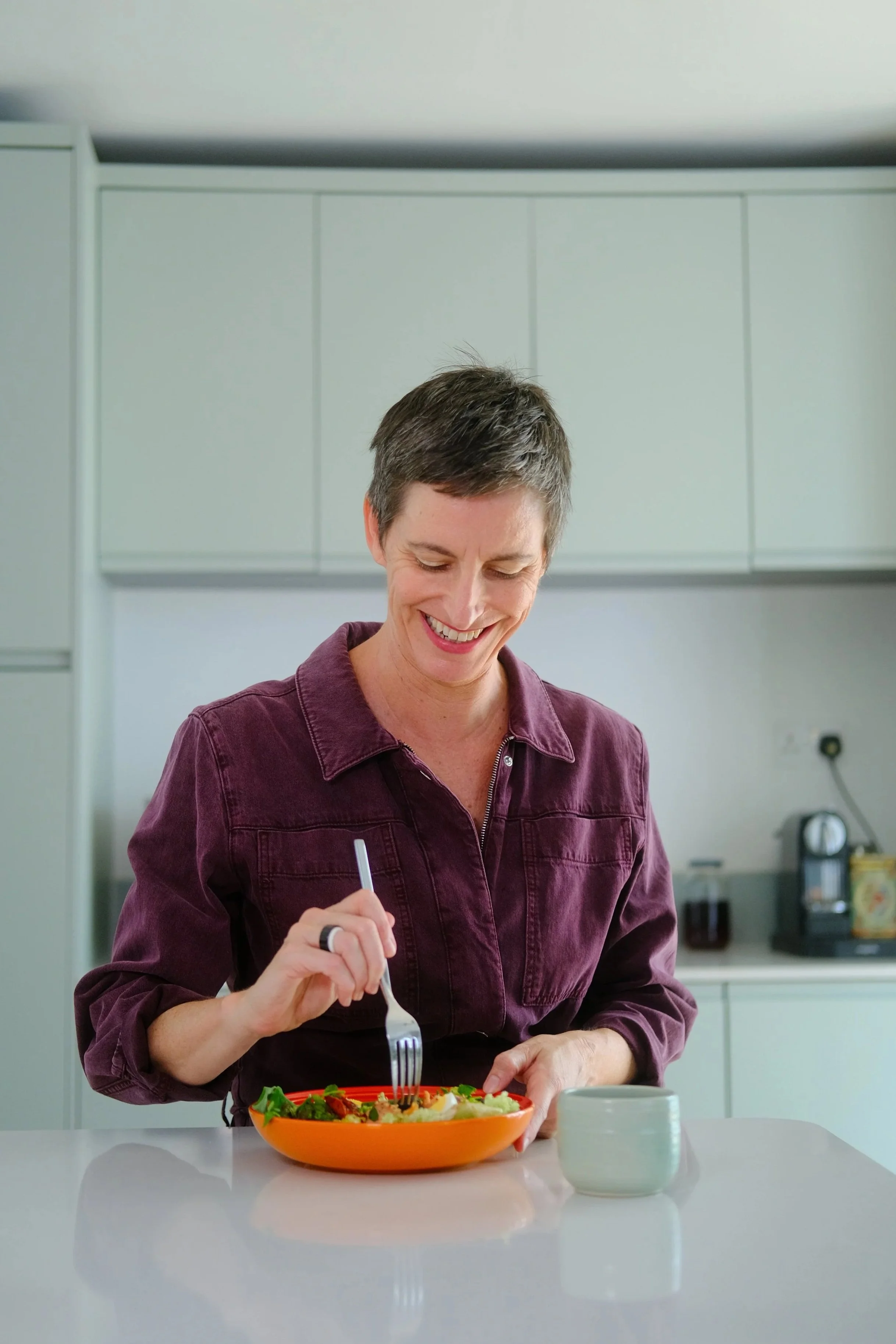 A smiling woman with short dark hair, wearing a maroon jumpsuit, sitting at a kitchen counter with a plate of salad in front of her. She holds a fork and is about to eat. A green mug is on the counter, and kitchen cabinets are in the background.