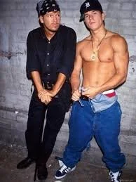 Two young men standing against a gray wall, one shirtless wearing baggy blue pants and a gold chain, the other dressed in a black shirt and black pants, wearing a bandana.