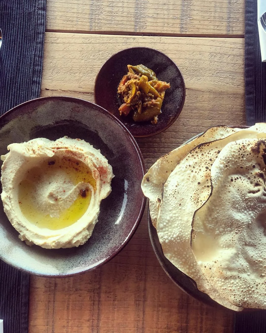 A bowl of hummus drizzled with olive oil, a small dish of cooked vegetables, and a basket of crispy, flatbread crackers on a wooden table.