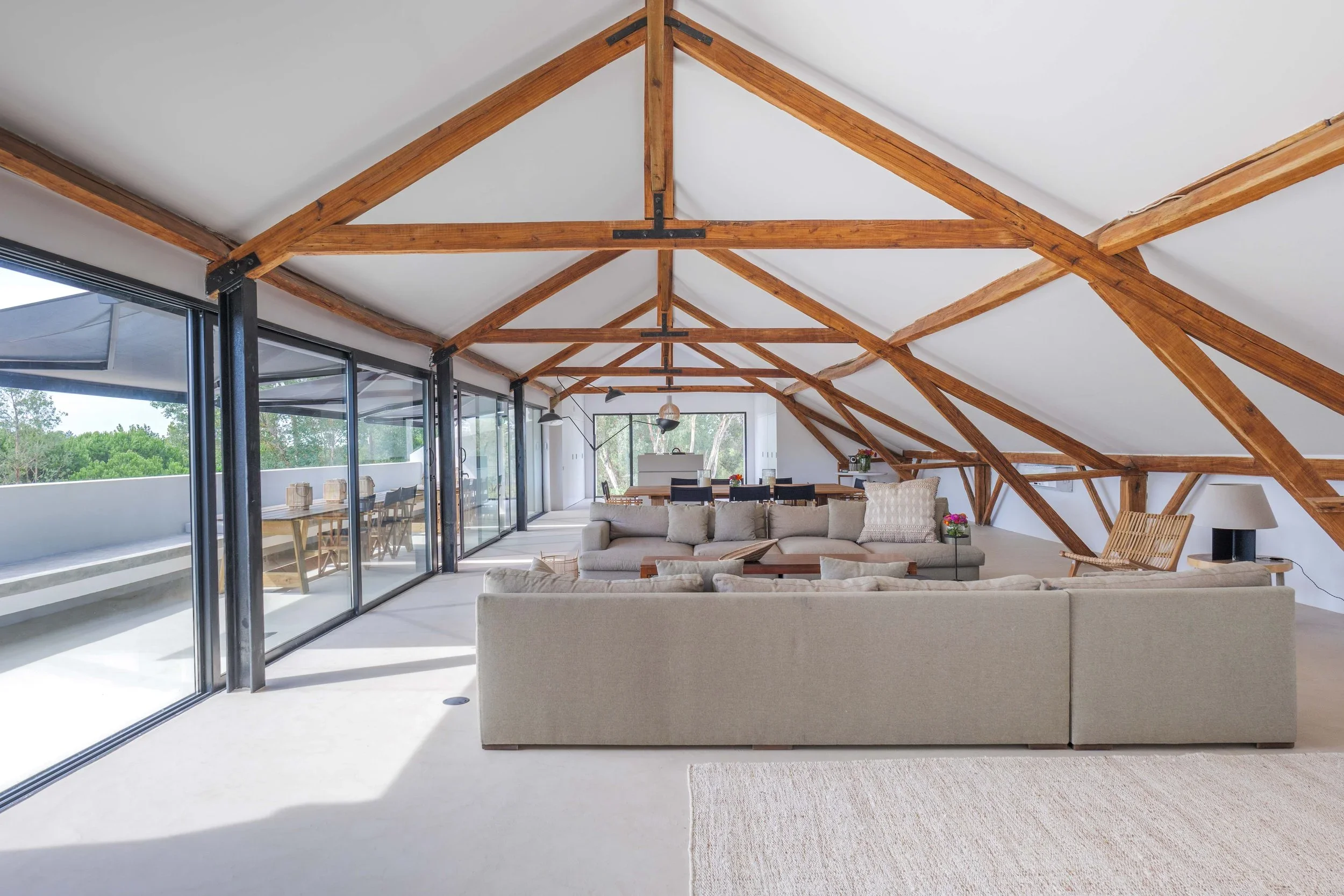 Spacious attic living room with white walls, exposed wooden beams, large glass sliding doors leading to balcony, beige sectional sofa, dining table with black chairs, and floor lamps.