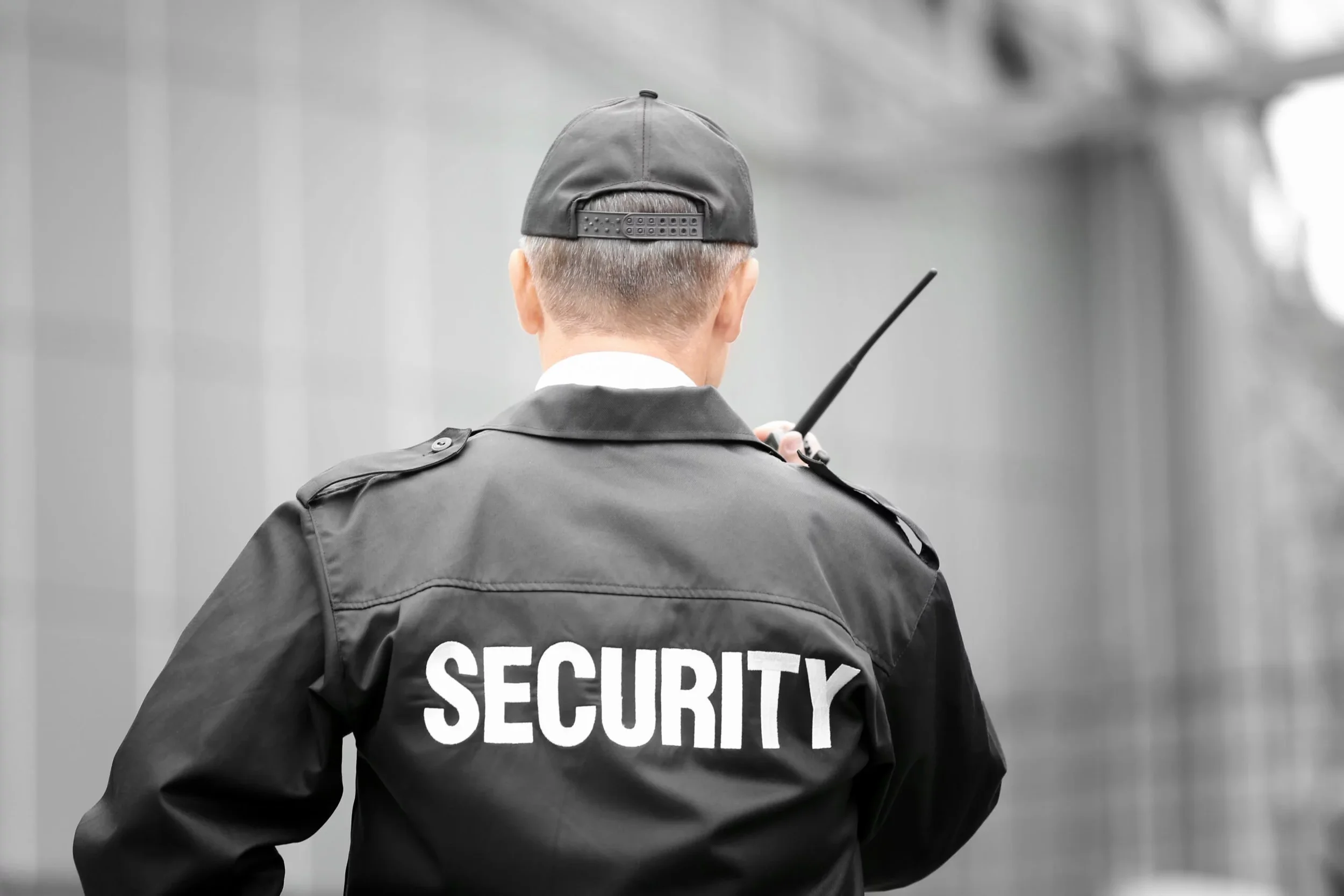 Security guard standing with back facing the camera, wearing a black jacket with 'SECURITY' written on it, and holding a walkie-talkie.