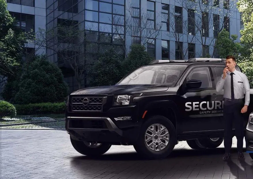 A security officer standing next to a black security vehicle talking on a mobile phone, with a modern office building and greenery in the background.