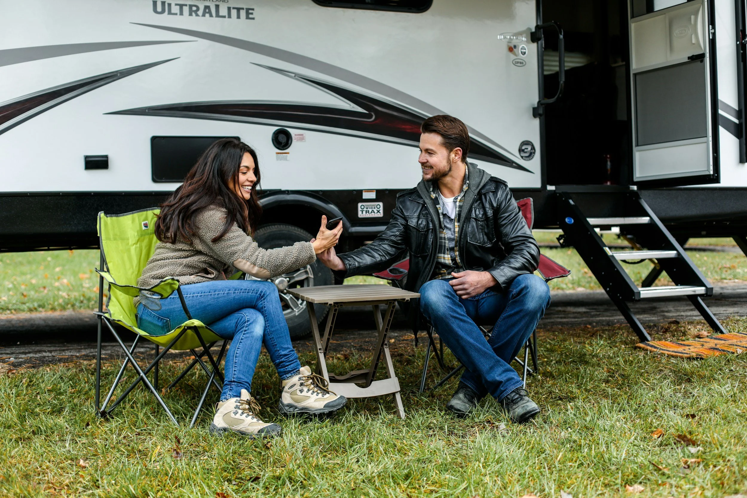 Un homme et une femme assis devant un camping-car en train de s'amuser, avec des chaises pliantes et une table. La femme sourit et semble prendre une photo ou un selfie avec le téléphone, l'homme lui tend la main. La scène se déroule en plein air, sur une aire verdoyante.