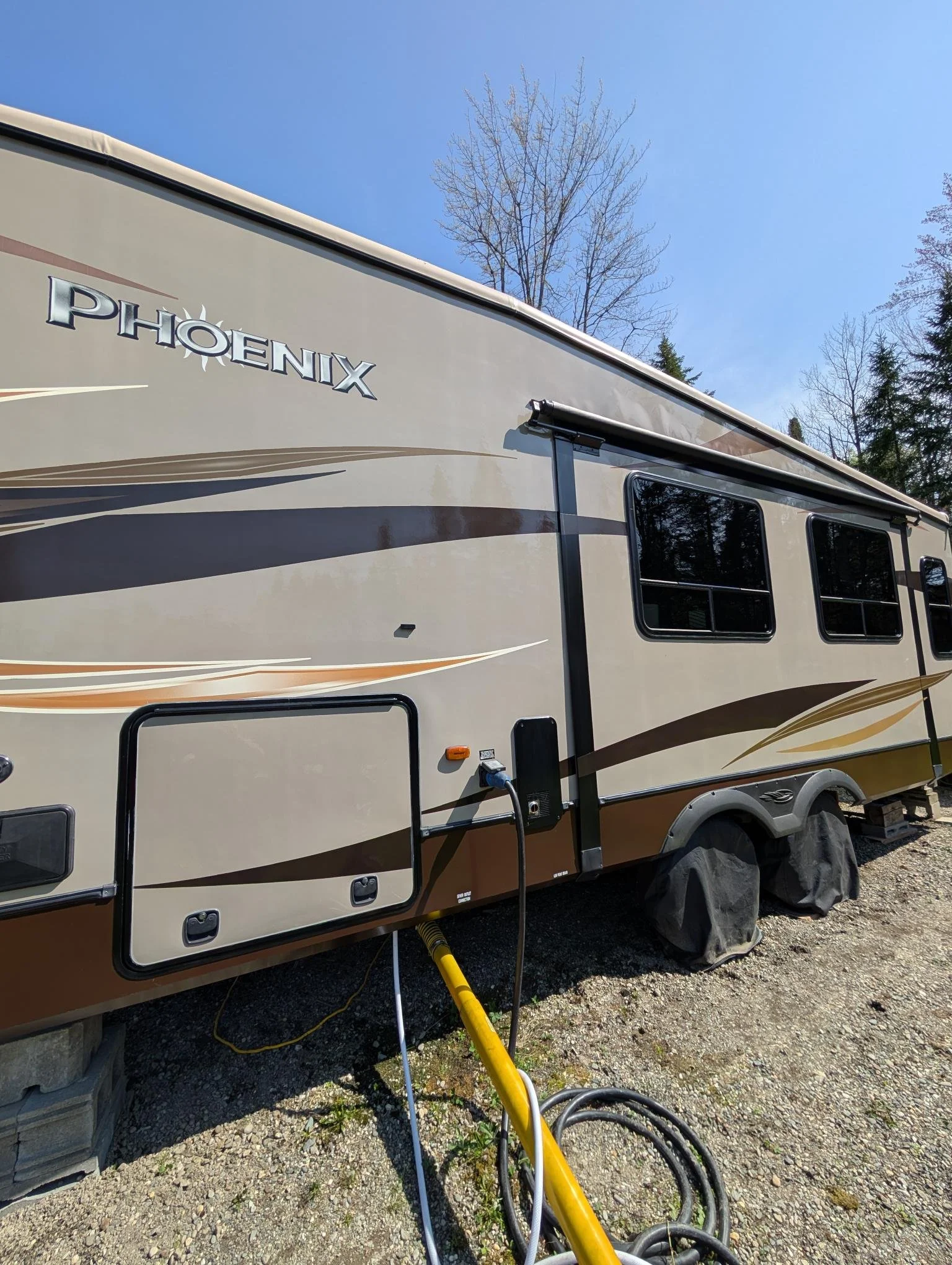 Camping-car beige avec des dessins décoratifs, stationné sur un terrain en pleine nature, avec des arbres en arrière-plan et un ciel bleu clair.