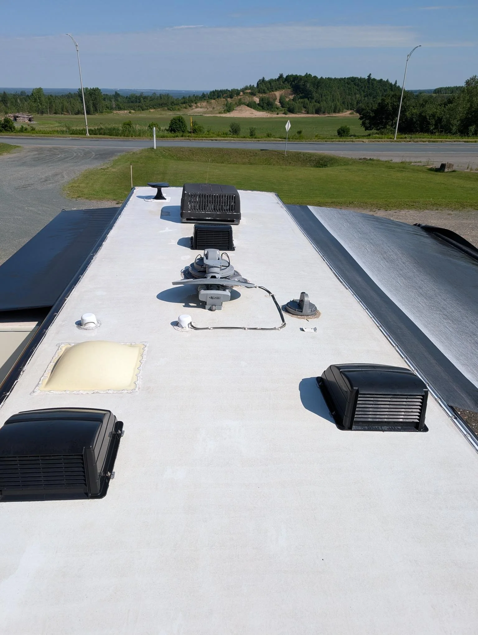 Vue de la toiture d'un camping-car avec ventilateurs et équipements, avec un paysage rural en arrière-plan sous un ciel bleu.