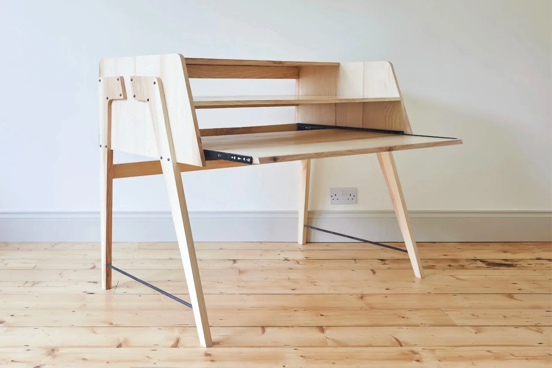 A  bespoke music production desk with shelves in a mid century style and made with ash pictured in a room with light wood flooring and white walls.