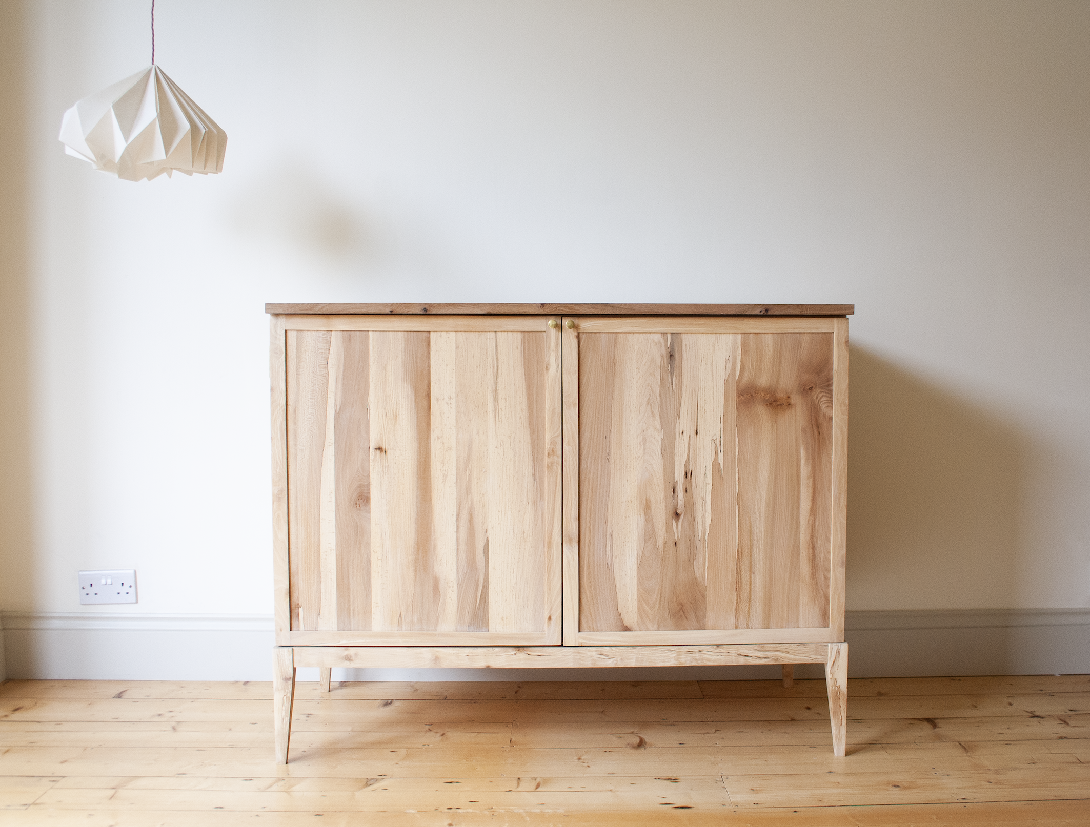 Light wood cabinet with two doors and gold knobs on a wooden floor, white wall background, and a white pendant light hanging from the ceiling.