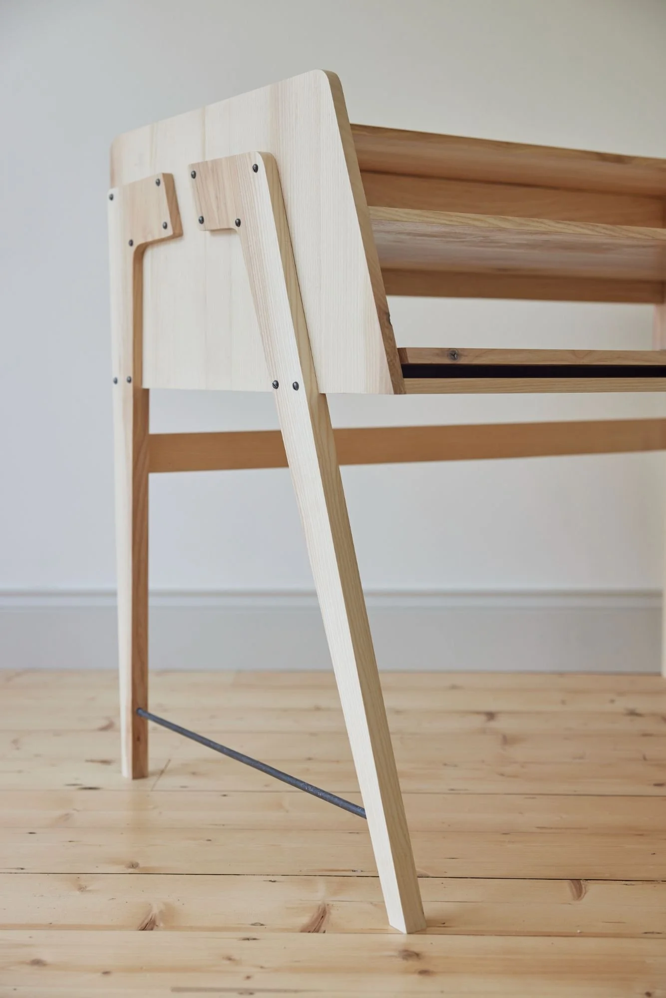 A minimalist wooden ladder with a natural finish leaning against a light-colored wall, with a wooden shelf partially visible behind it.