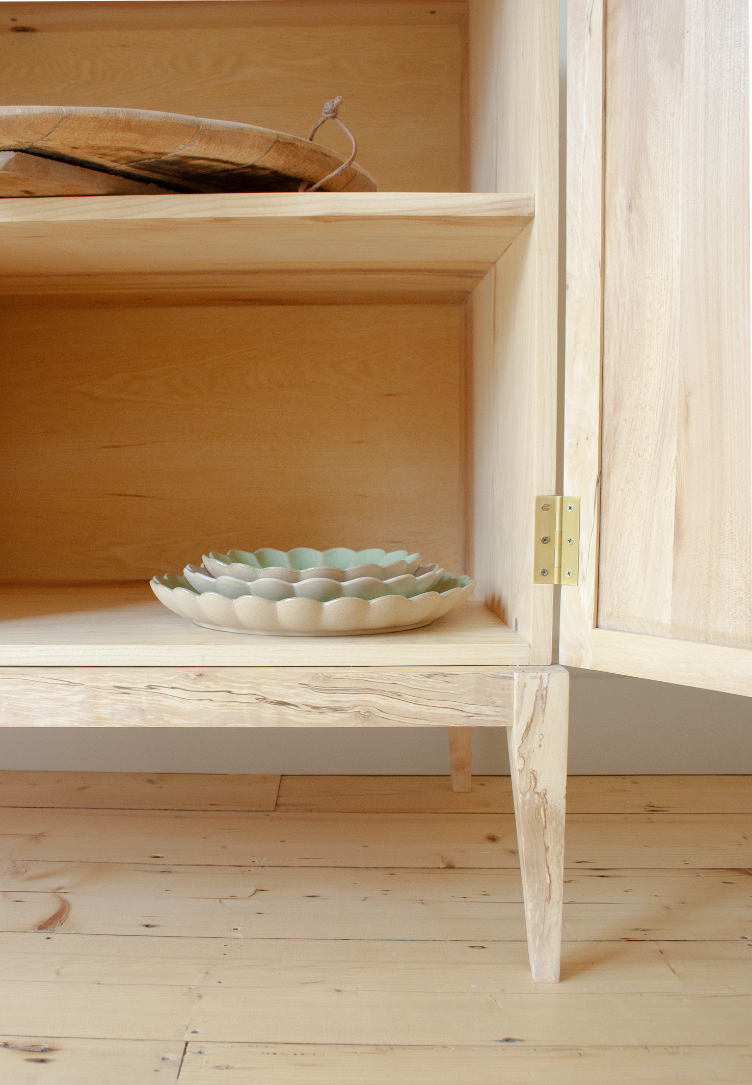 Open wooden cabinet with three ceramic plates on the bottom shelf, a wooden tray on the top shelf, and a wooden floor.