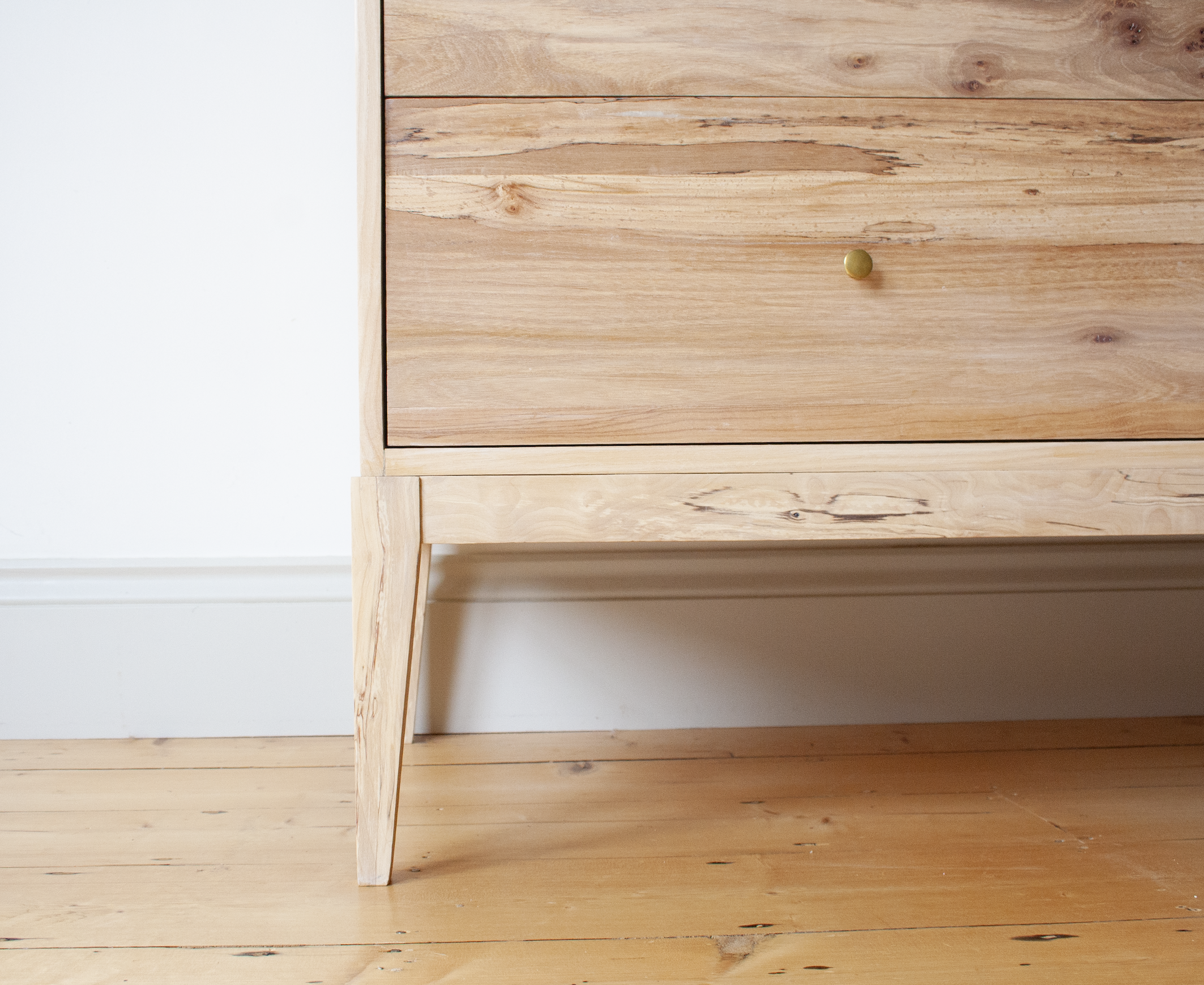 Close-up of a wooden dresser with a single brass-colored knob, standing on four angled legs on a wooden floor near a white wall.