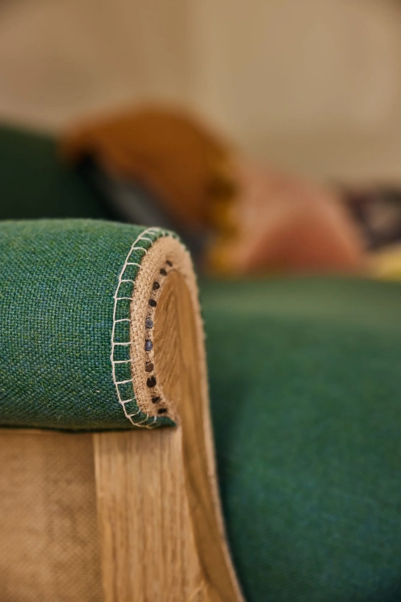 Close-up of a green upholstered furniture arm with white stitching, on a wooden frame, with a blurred background