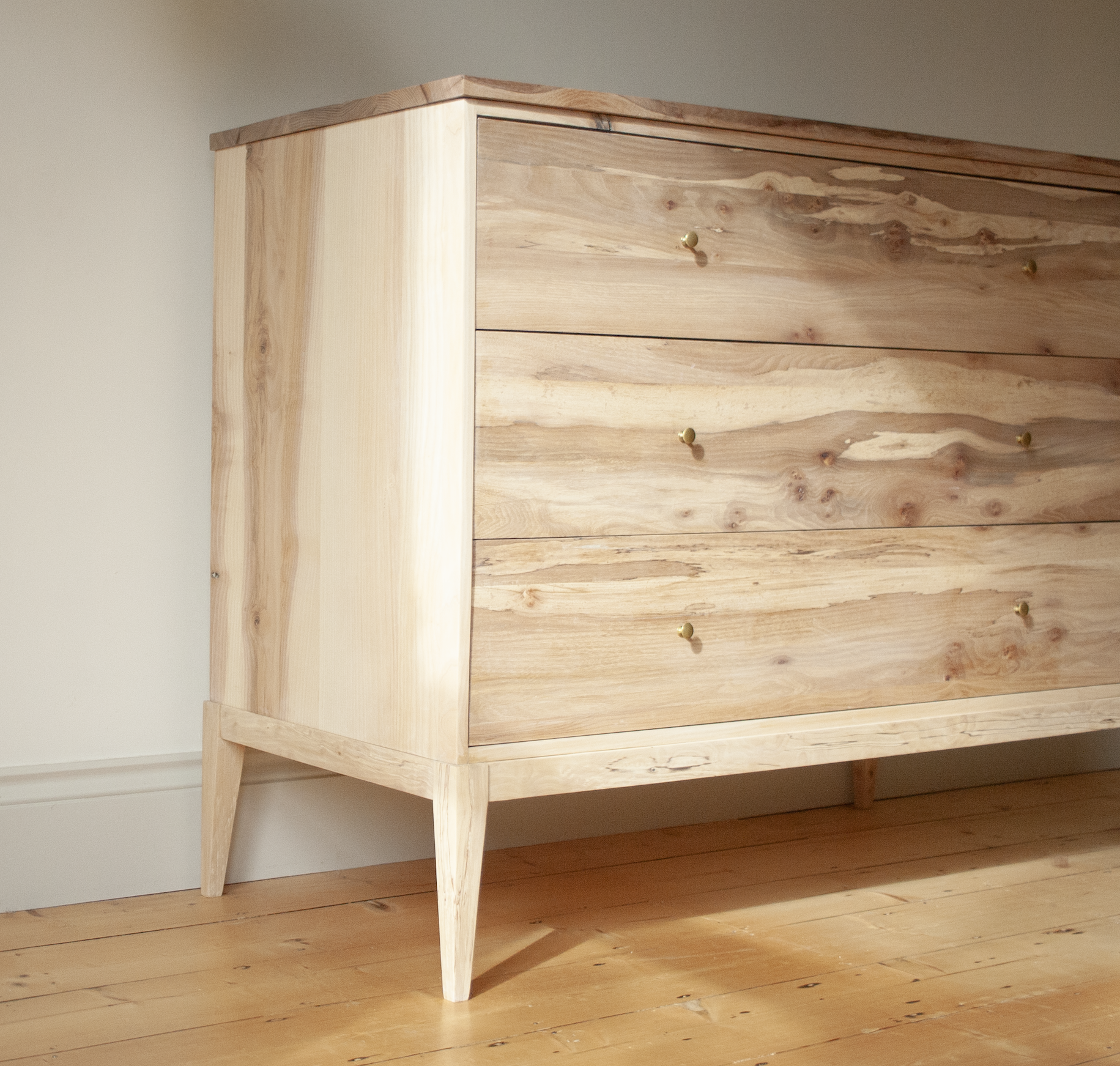 Light wood dresser with gold knobs on a wooden floor, against a white wall.
