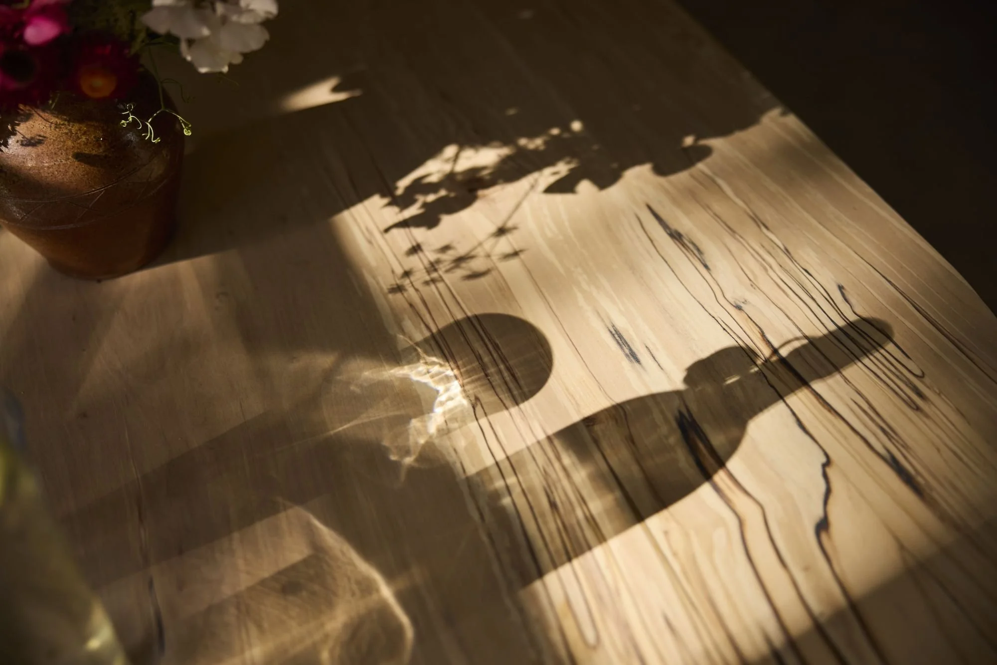 Shadows of a vase with flowers, a bottle, and a small object cast on a wooden table top made with solid silver birch by Trew & Bold
