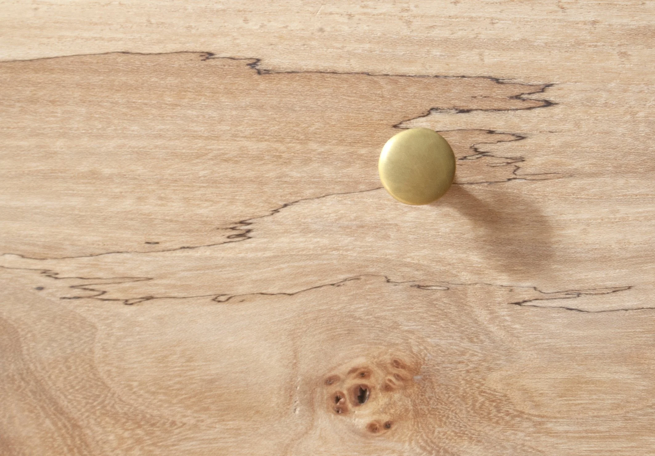 Close-up of a wooden surface with natural knots and grain patterns, featuring a single round, smooth, gold-colored object casting a shadow.