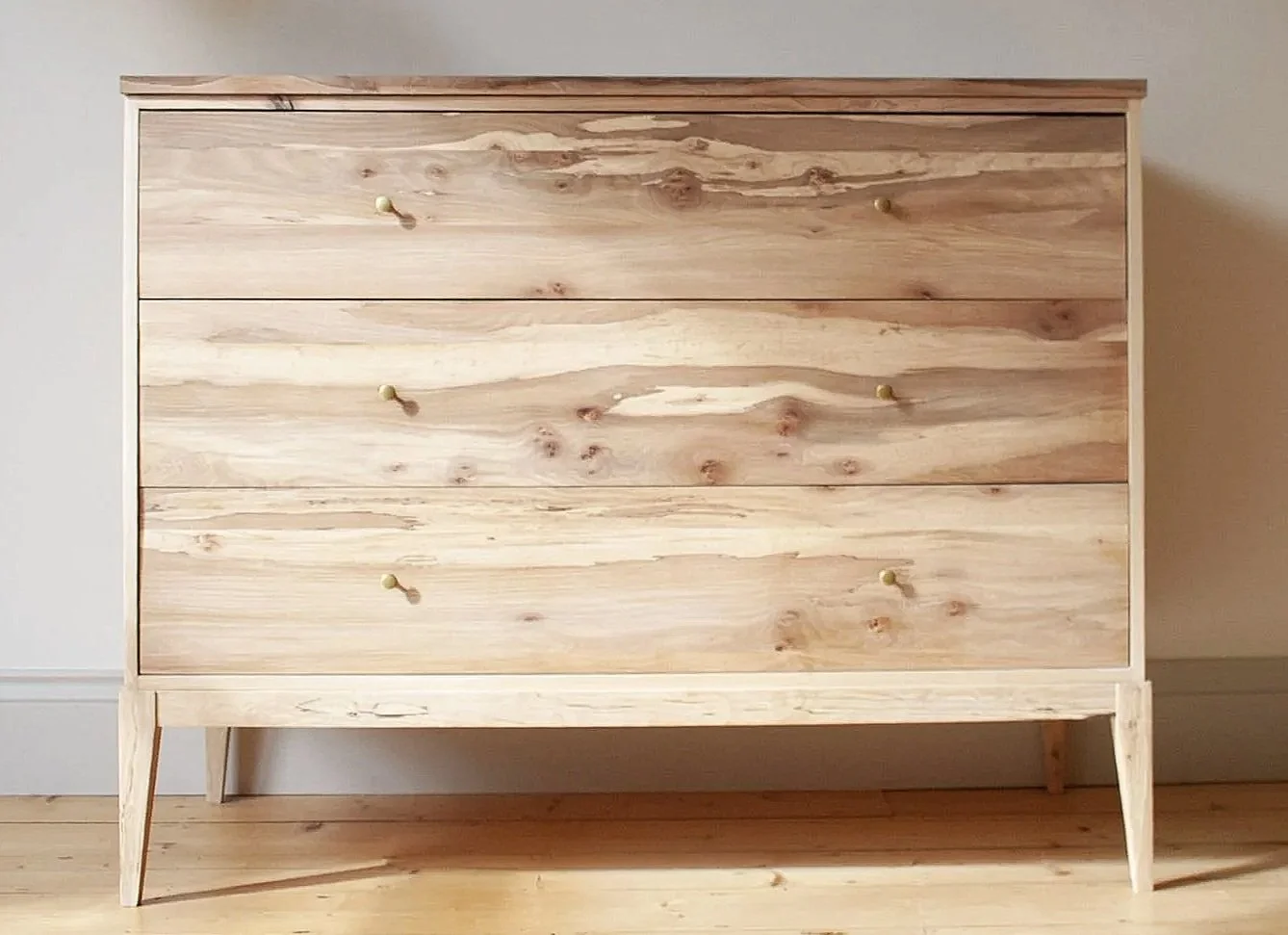A solid wood chest of drawers in a minimal design made from an ash carcase with spalted elm drawer fronts and brass drawer pulls