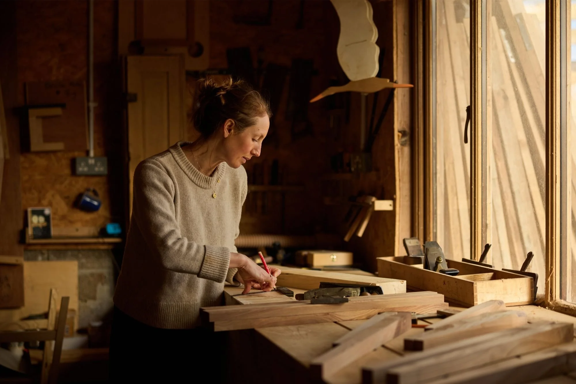 Emily, Trew and Bold founder working in a woodworking shop, bent over a workbench, drawing with a red pencil on a wooden board, surrounded by tools, with warm lighting highlighting her and the wood.