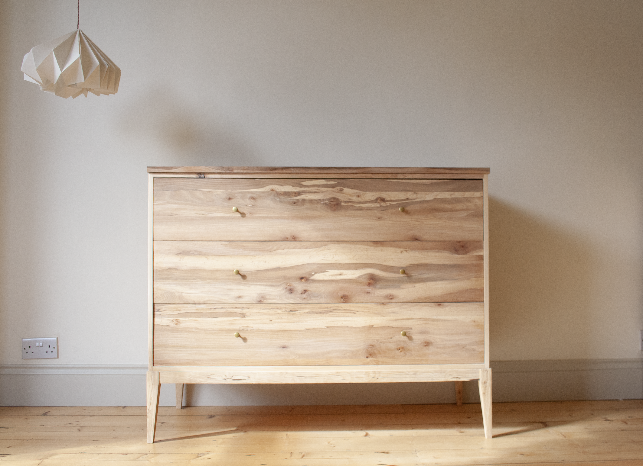 A wooden dresser with three drawers and round knobs, standing against a plain beige wall in a room with wooden flooring and a paper pendant lamp hanging from the ceiling.