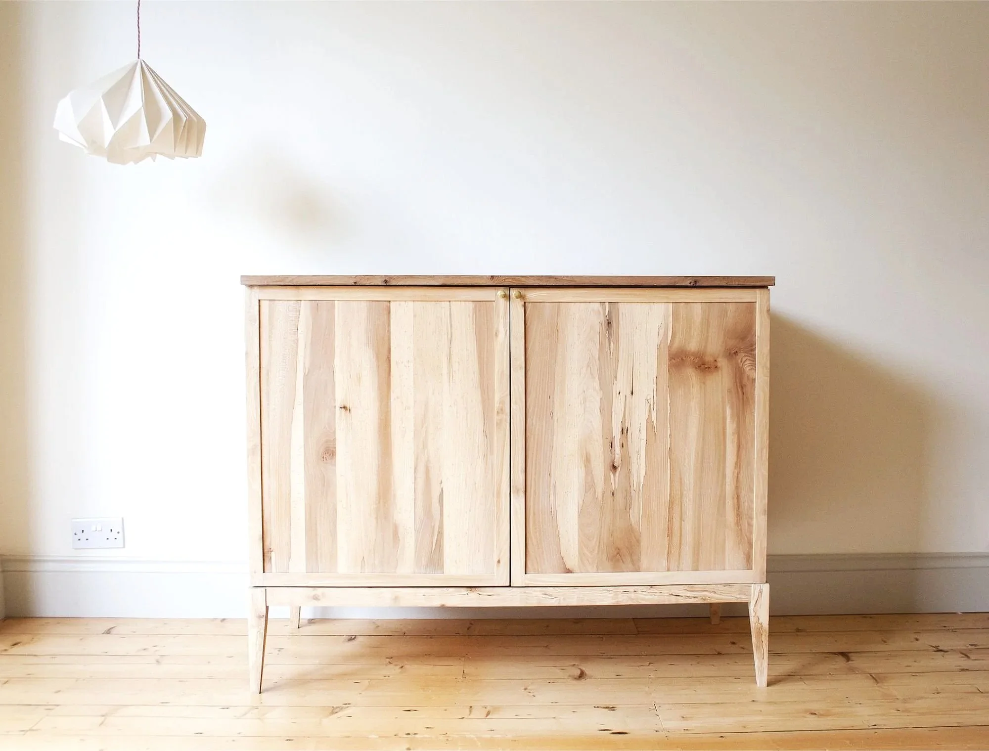 A shaker style cabinet with slim shaker doors, elegant tapered legs and spalted elm door fronts standing in a room with light-colored walls and wooden flooring, near a wall socket and electrical outlet.