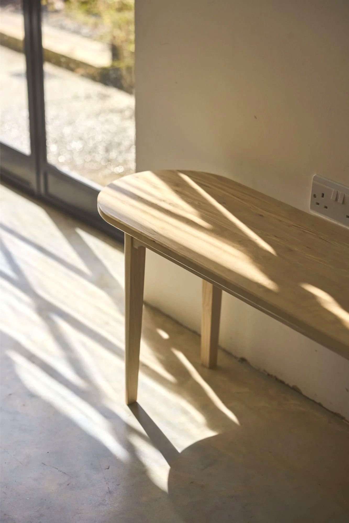 A wooden bench with a rounded edge positioned on a concrete floor next to a wall with a power outlet. Sunlight casts shadows on the floor and table, with a large glass door or window in the background showing an outdoor area.
