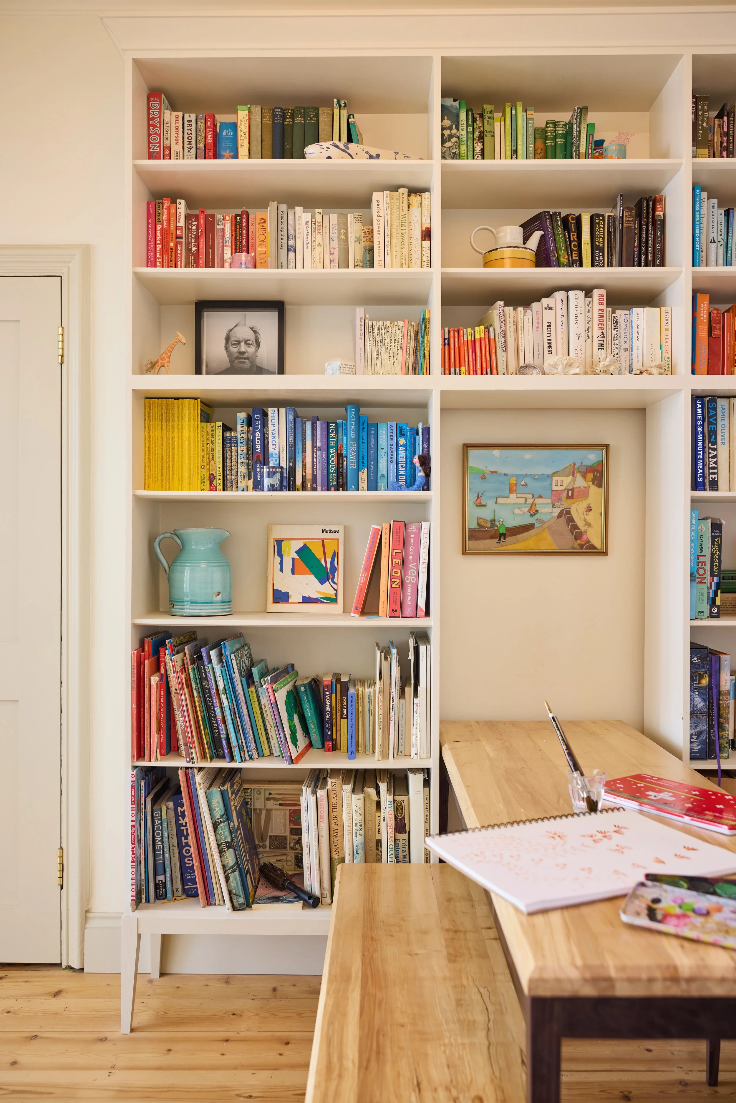 White bookshelf filled with books, a framed black and white photo, a green vase, and a colorful painting on the wall. A wooden table made with silver birch by furniture studio Trew and Bold with notebooks and art supplies is in front.