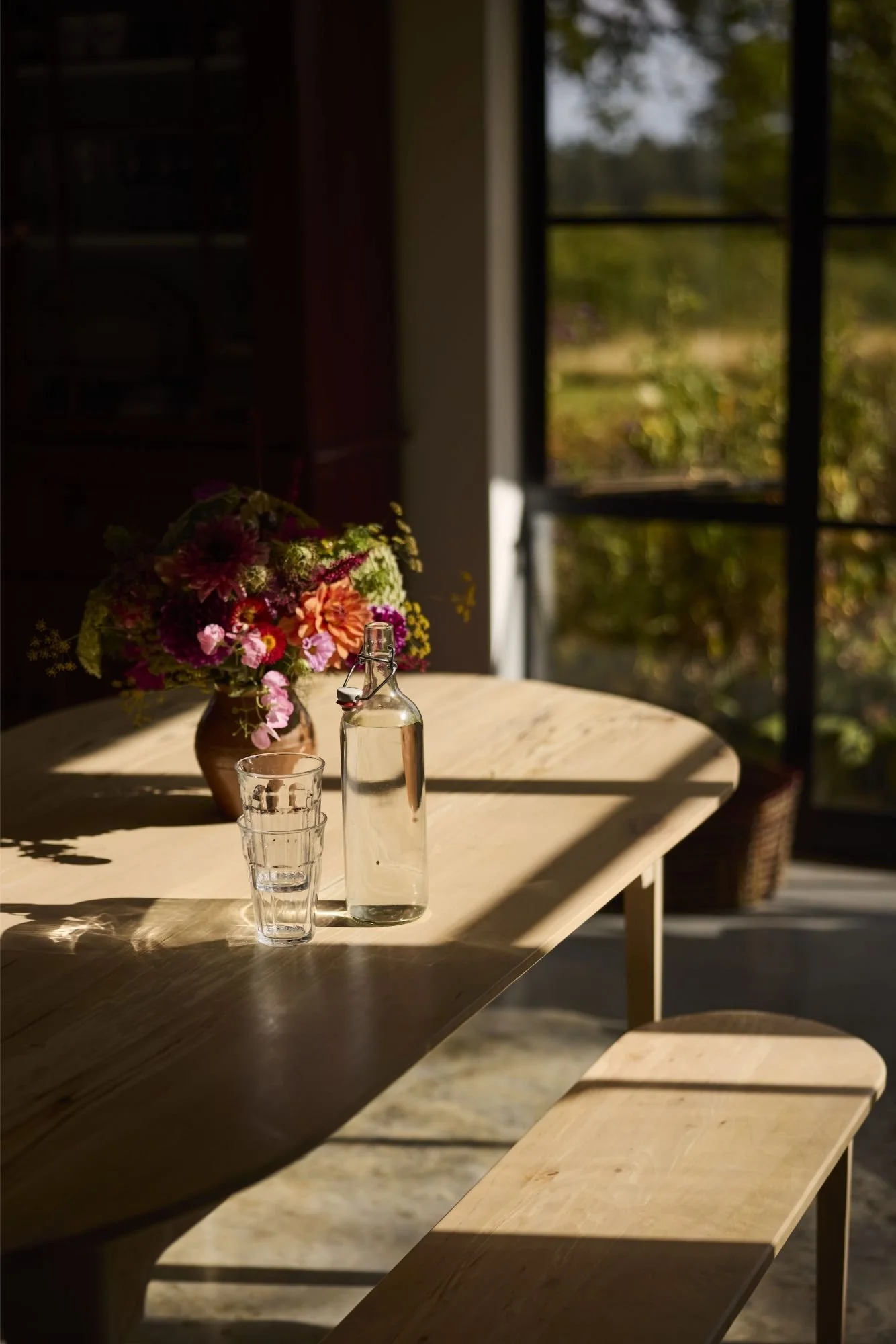 A wood dining table and bench set with sunshine casting beautiful shadows across the top and colourful flowers
