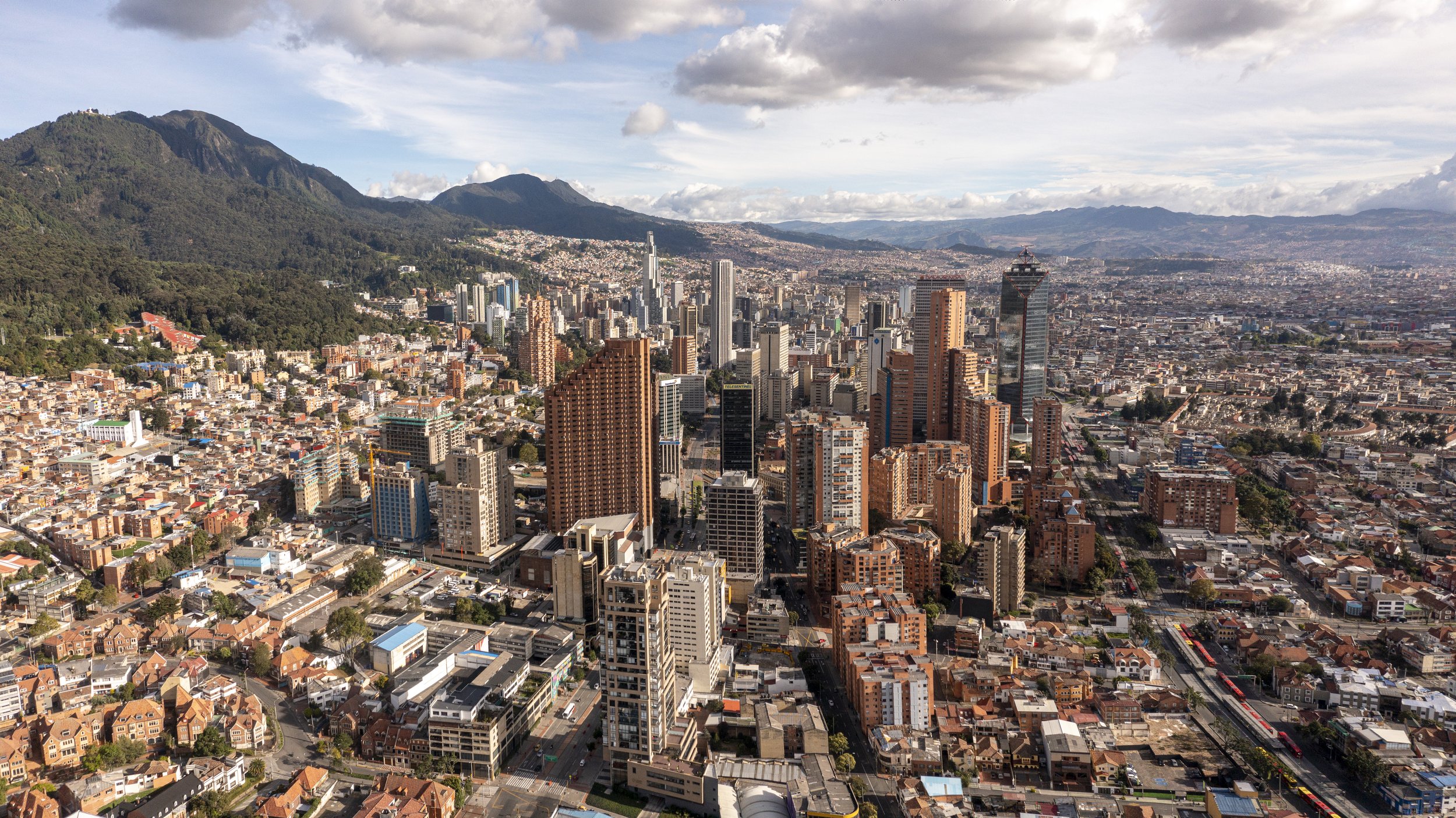 A city skyline with tall skyscrapers, surrounded by mountains in the background, under a partly cloudy sky.