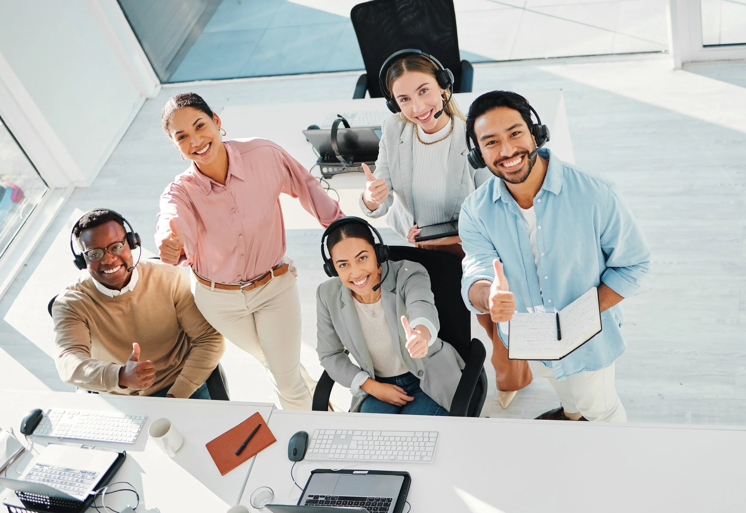 Five diverse office workers wearing headsets and giving thumbs up, gathered around a desk with computers and notebooks, in a bright work environment.