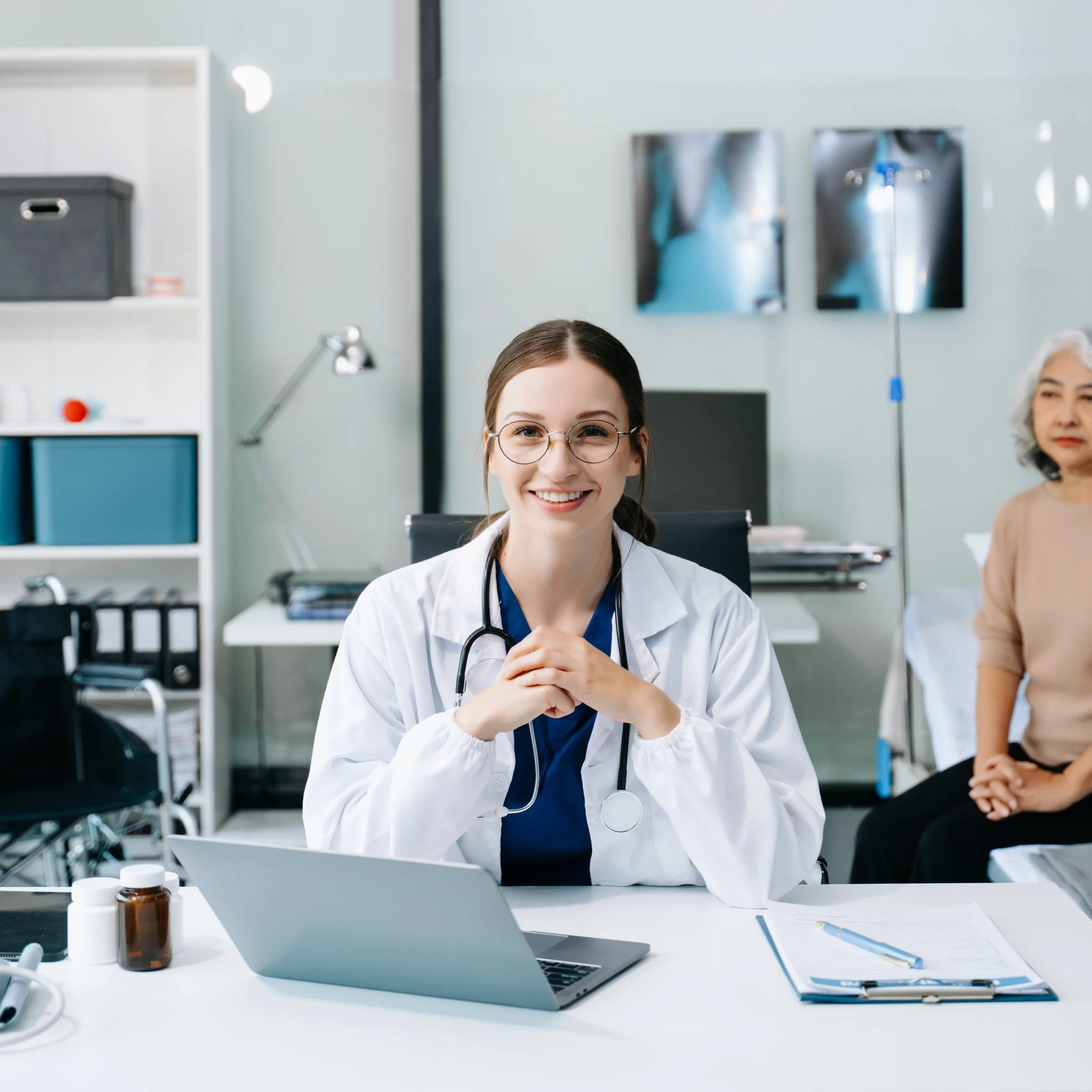 A doctor in a white coat with a stethoscope sits smiling at a desk in a medical office, with a laptop, documents, and medication bottles. In the background, a patient sits on an examination table. The room has medical equipment and shelves with files.