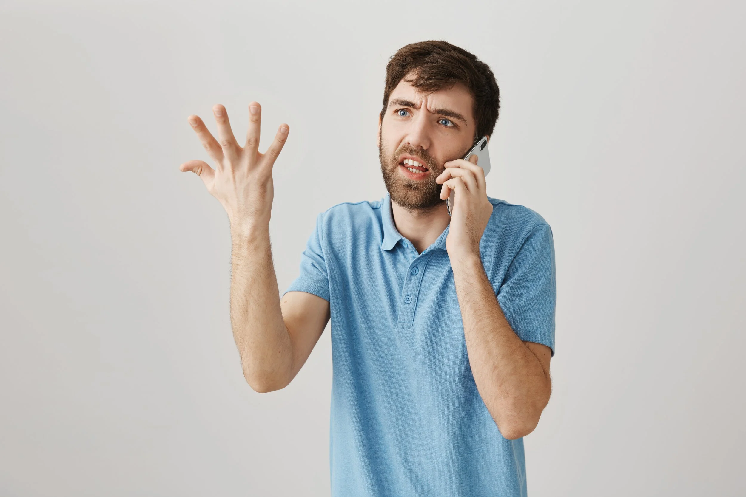 A confused young man with brown hair and beard, wearing a blue polo shirt, talking on a mobile phone, with his left hand raised and fingers spread, expressing confusion or frustration.