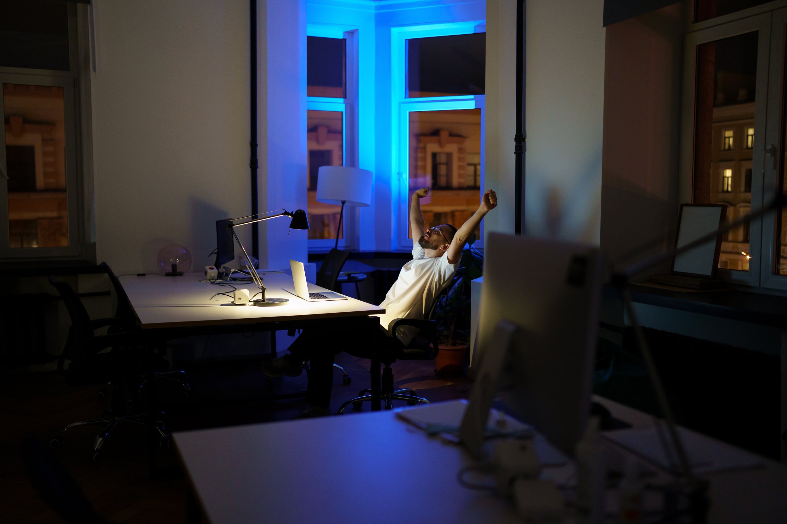 A man sitting in an office chair at a cluttered desk, stretching with arms raised behind his head, in a dimly lit office at night with blue and yellow lighting outside the windows.