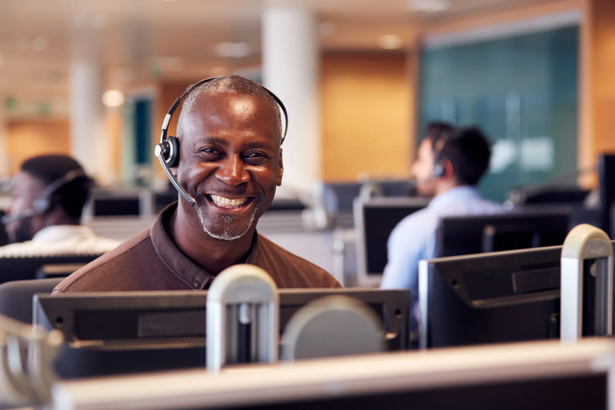 A smiling man wearing a headset sits at a desk in a call center with other agents working in the background.
