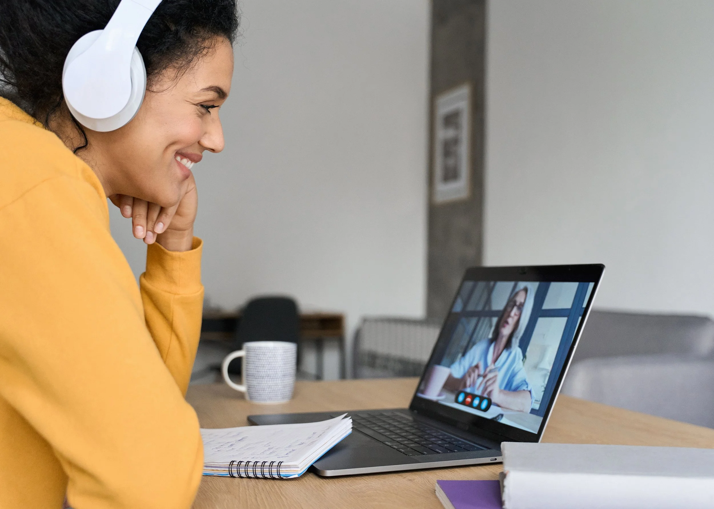 Woman in yellow sweater wearing white headphones participates in video call on laptop, taking notes in a notebook on a wooden table.