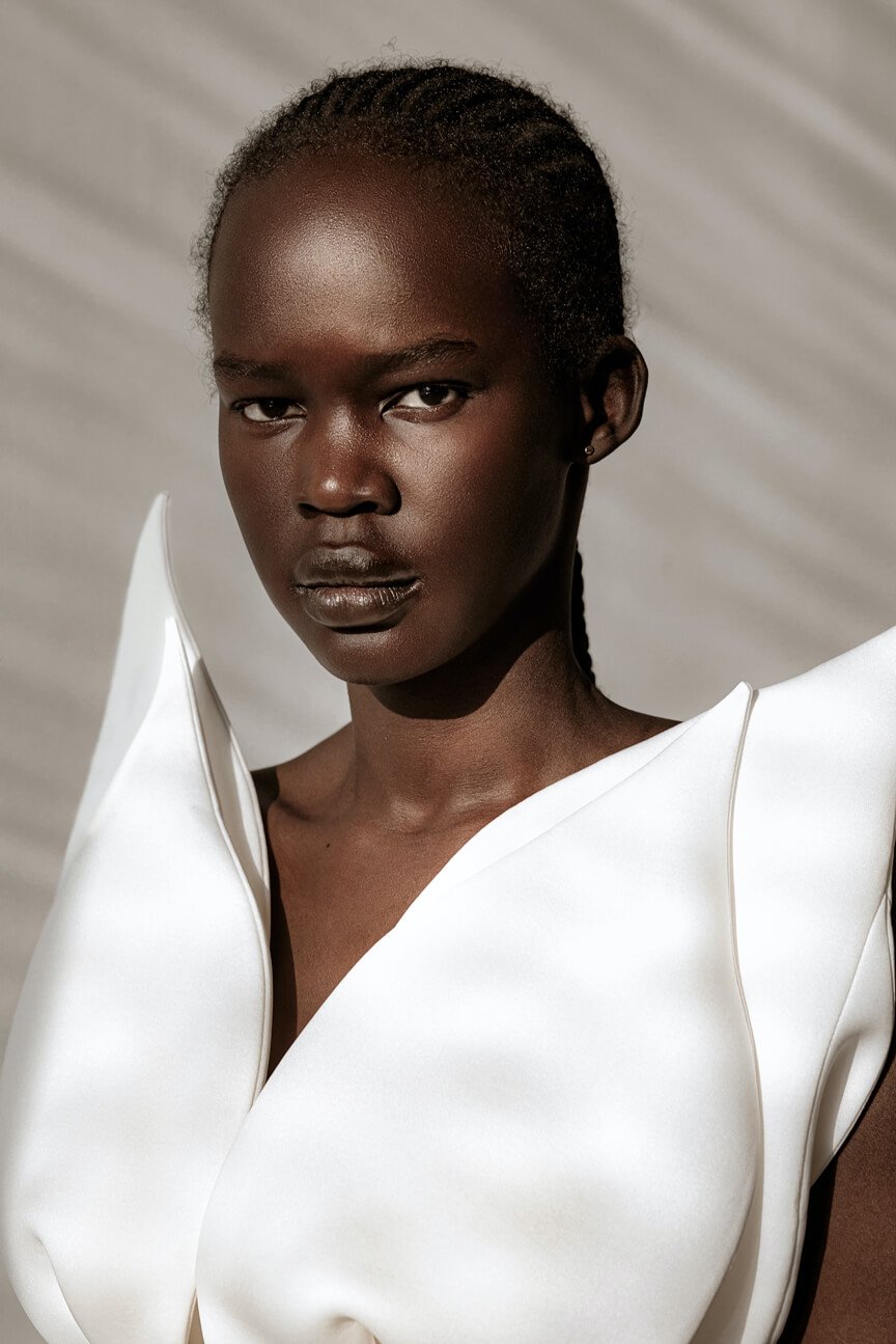 A woman with dark skin and short hair styled in braids, wearing a white top with structured shoulders, looking directly at the camera. The background is neutral and simple.