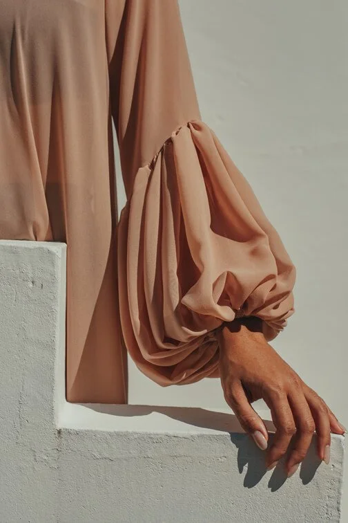 Close-up of a person's hand hanging over a white ledge, wearing a beige long-sleeve shirt with large, puffed sleeve design.