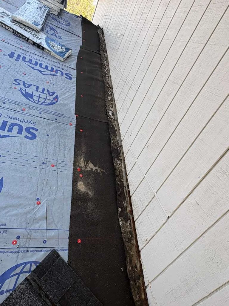 Close-up of a residential roof edge showing waterproofing membrane and underlayment with flashing, vinyl siding on the exterior wall, and roofing nails.
