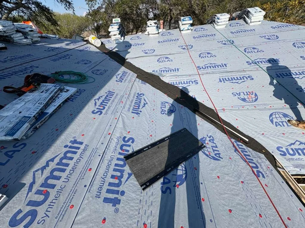 Construction site with roofing underlayment, tools, bundled materials, and a worker's shadow, with trees in the background.