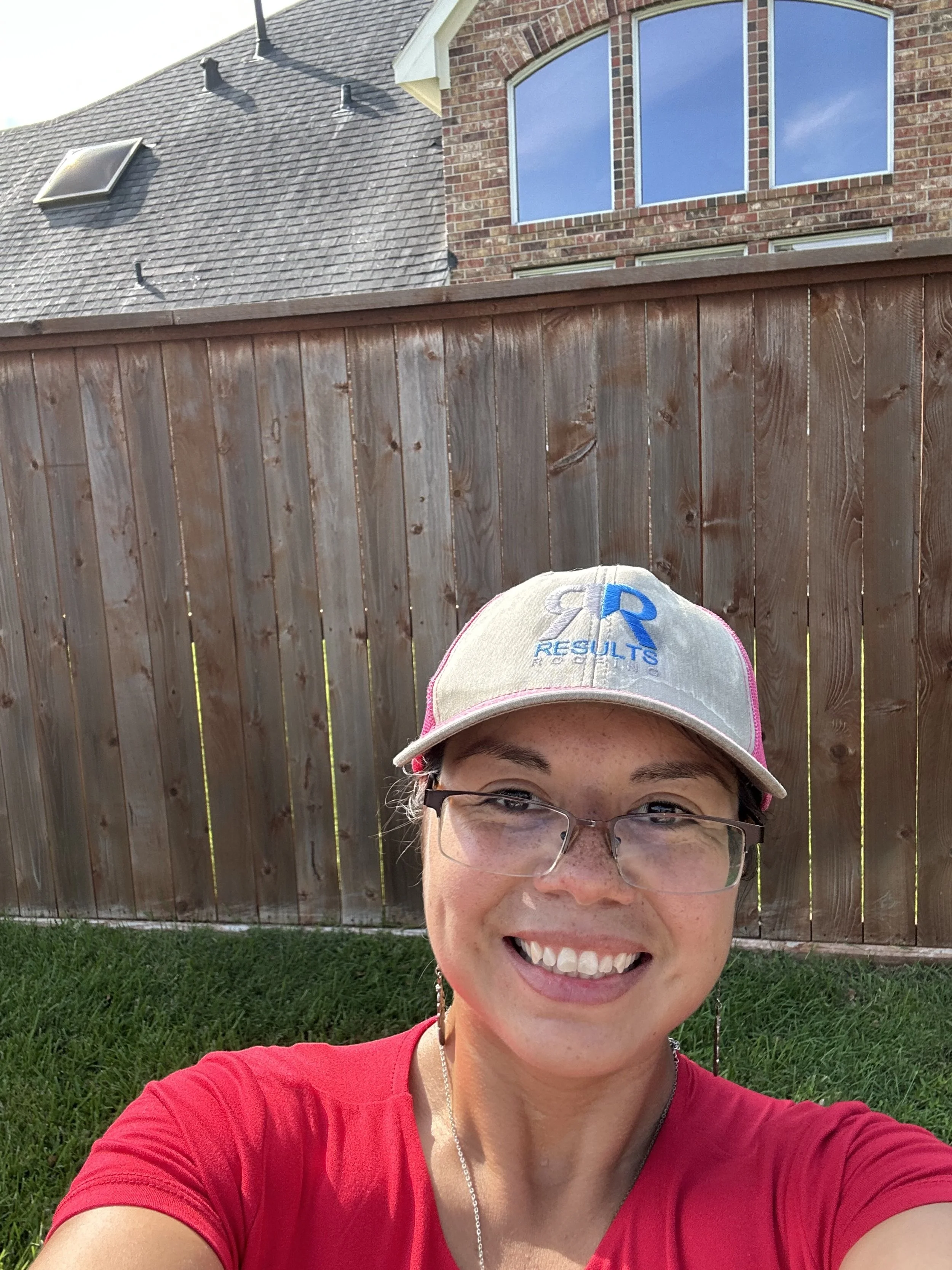 A woman smiling outdoors in front of a wooden fence with a house and window in the background.