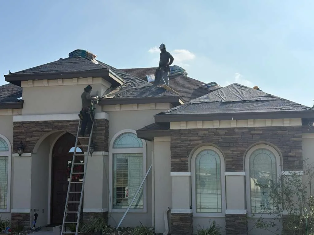 Two workers working on roof after being felted over on a large, beige house with arched windows, using a ladder and tied-off safety equipment.