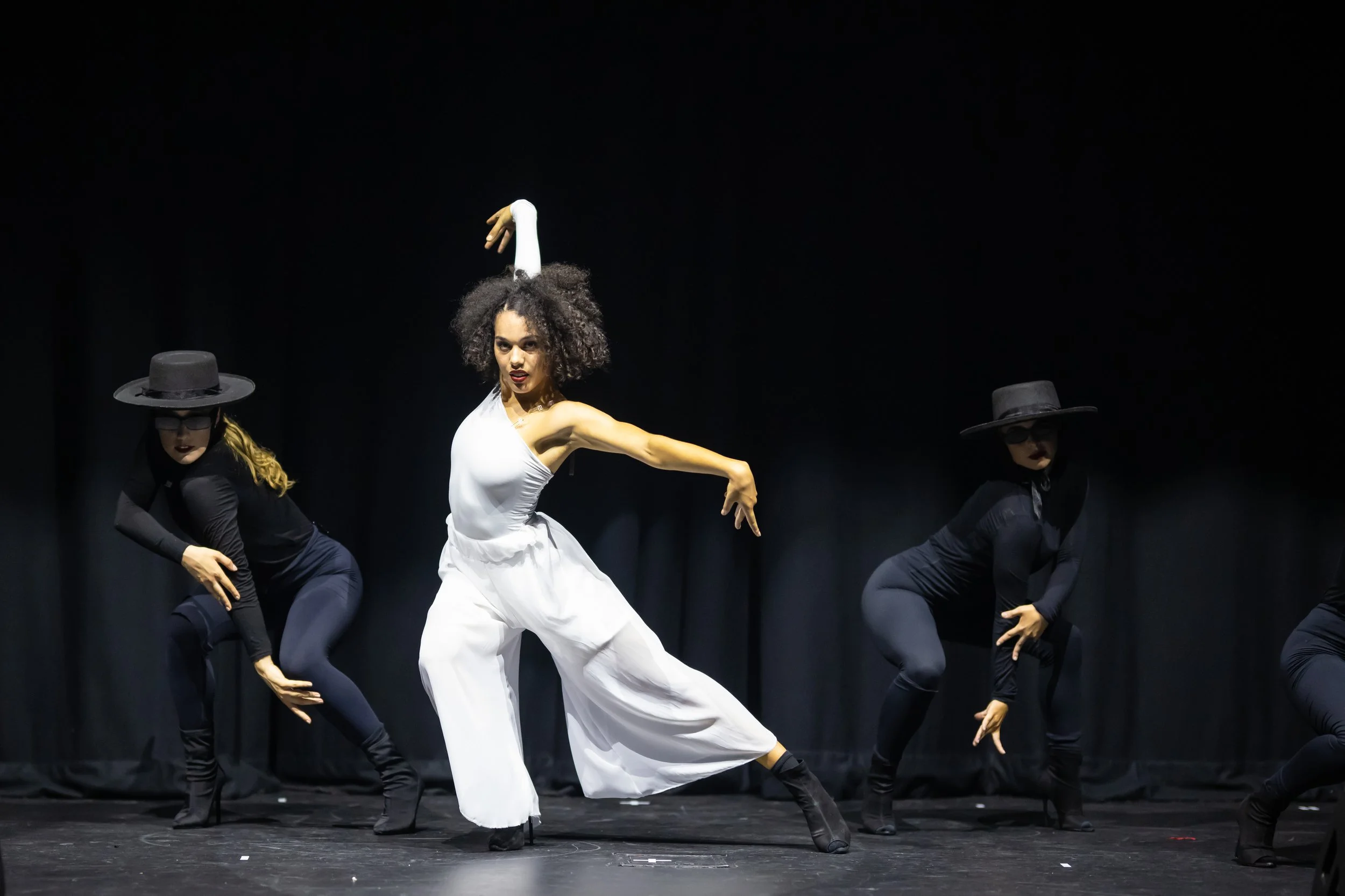A woman in white jumpsuit and two women in black outfits and wide-brimmed hats perform a dance on stage with a black background.