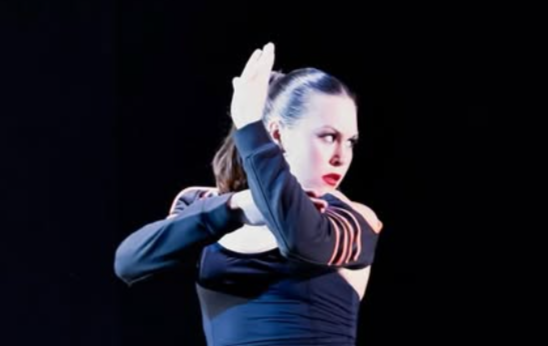 Dancer with braided hair and red lipstick performs ballet or contemporary dance on stage against a black background.