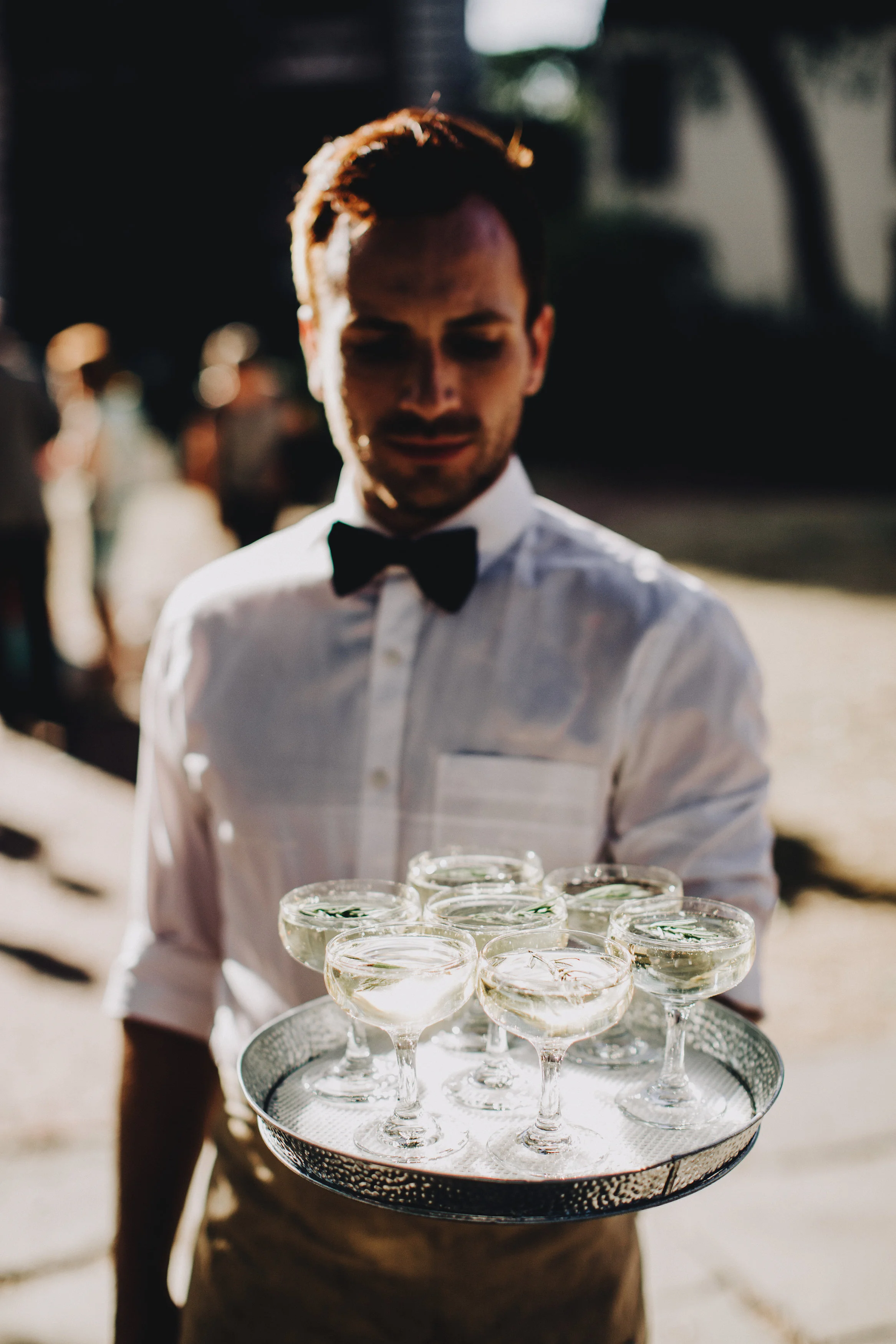 A bartender in a white shirt and black bow tie holding a tray of cocktails outdoors.