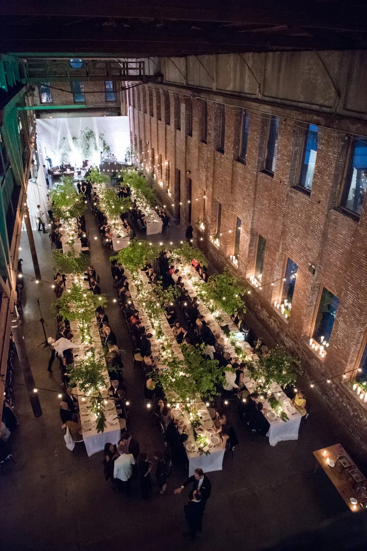 An elegant indoor wedding reception with long tables decorated with greenery and candles, hanging string lights, and guests seated under a brick wall backdrop.